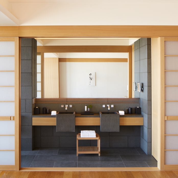 Warm blonde wood frames dual vanities in black stone, where traditional Japanese screens slide open to reveal a serene soaking sanctuary.