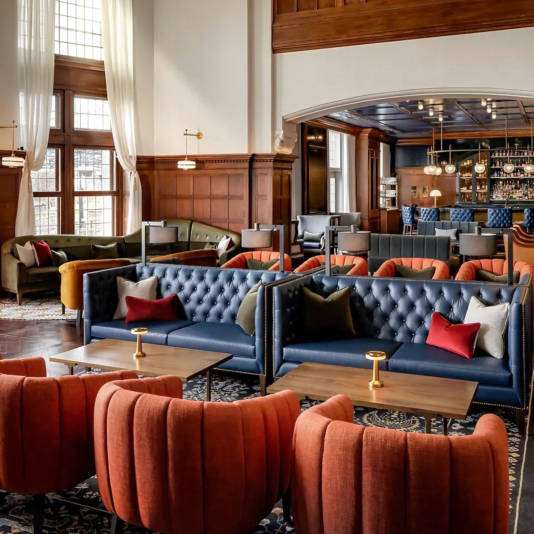 Rich navy leather banquettes and burnt orange armchairs create intimate conversation nooks beneath soaring ceilings, where golden light filters through tall windows onto polished wood paneling.
