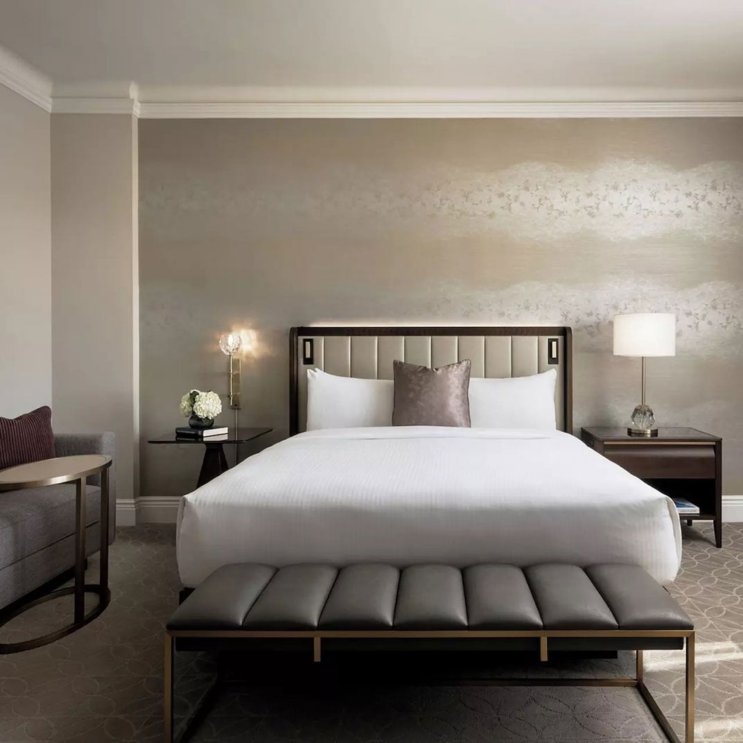 Champagne-hued wallpaper creates luminous bands of light above crisp white linens, while polished nightstands and a tufted bench add sophisticated warmth to this refined guest room.