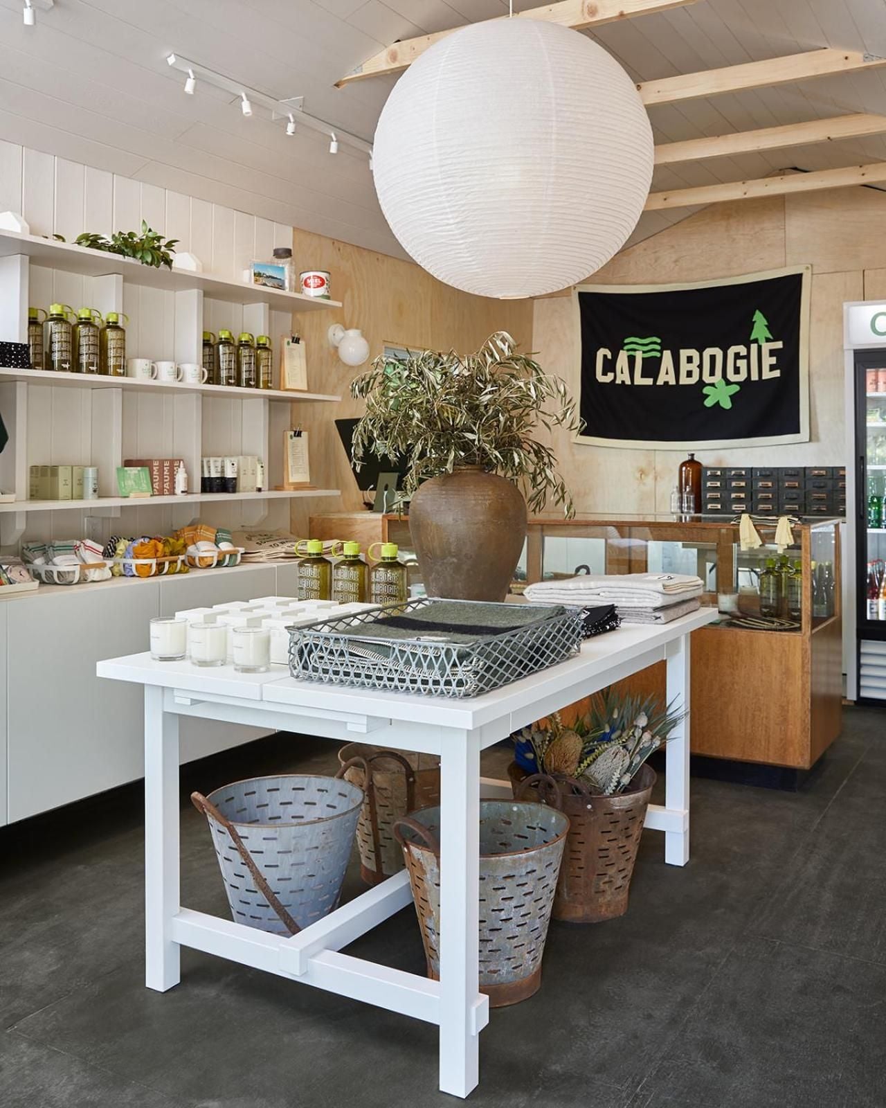 Curated home goods and local provisions fill whitewashed shelves beneath exposed beams, while wicker baskets and handmade ceramics create an inviting marketplace atmosphere.