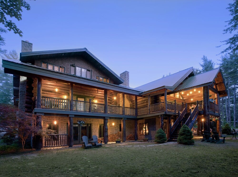 Warm light spills from timber balconies as twilight settles over this handcrafted wilderness retreat, where rustic logs meet refined comfort in Nova Scotia's forest.