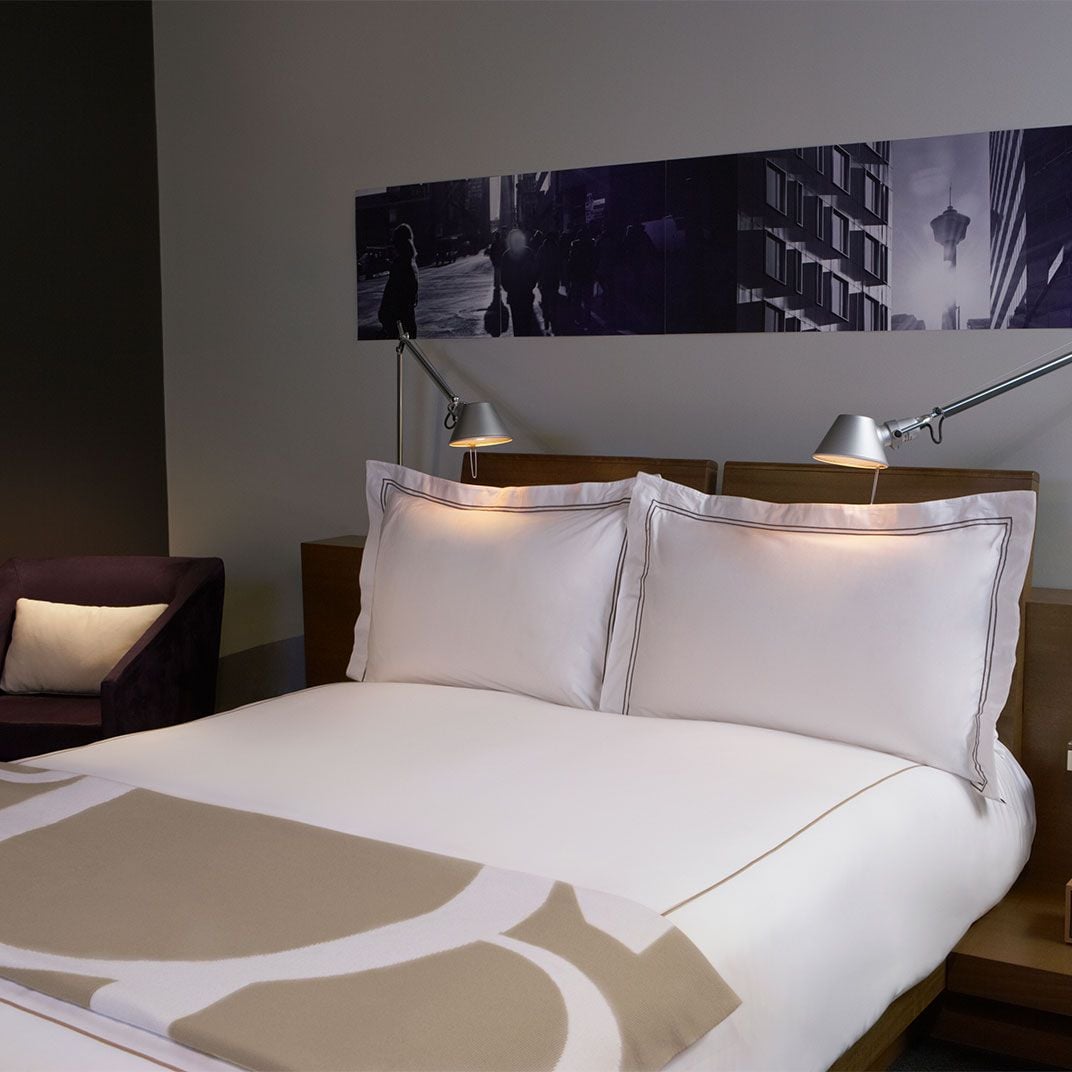 Crisp white linens with tailored brown piping create a sophisticated sleep sanctuary, while adjustable reading lamps cast warm pools of light against the urban photography above.