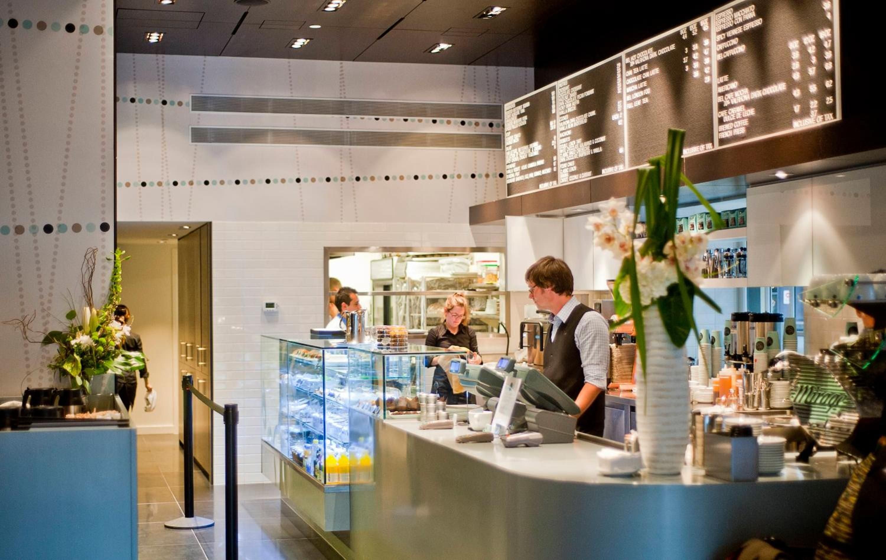 Bright modern café buzzes with Vancouver energy as baristas craft artisanal drinks beneath illuminated menu boards and contemporary dotted wall art.
