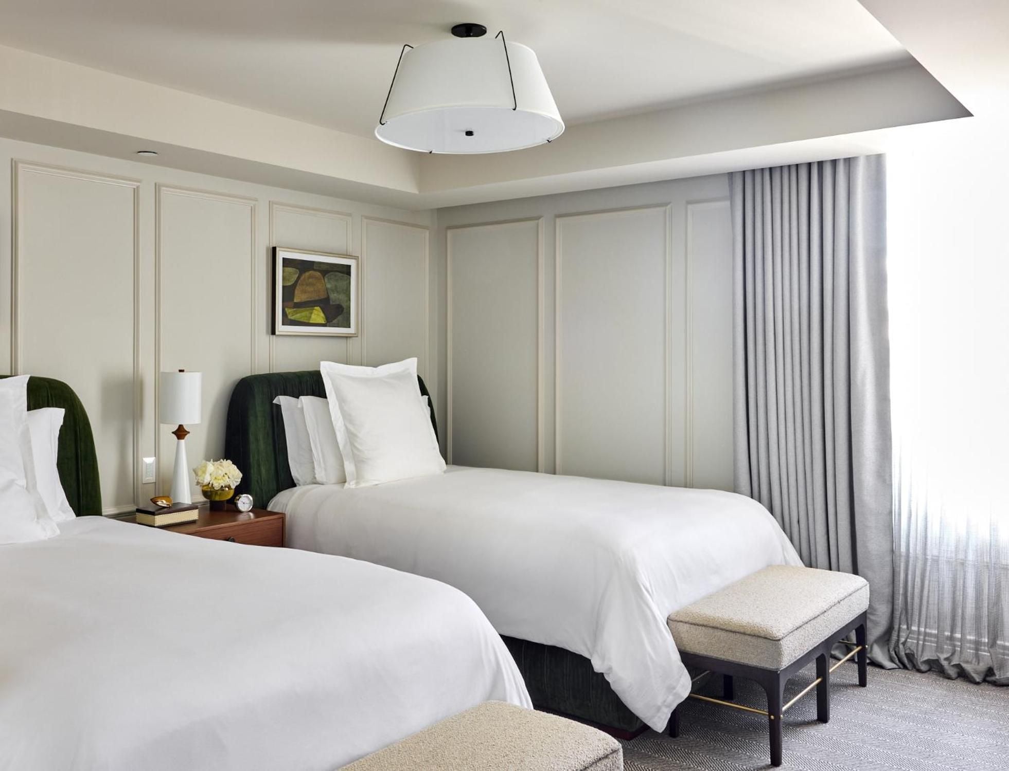 Crisp white linens contrast beautifully with forest-green velvet headboards in this sophisticated twin bedroom, where paneled walls and soft gray curtains create timeless elegance.
