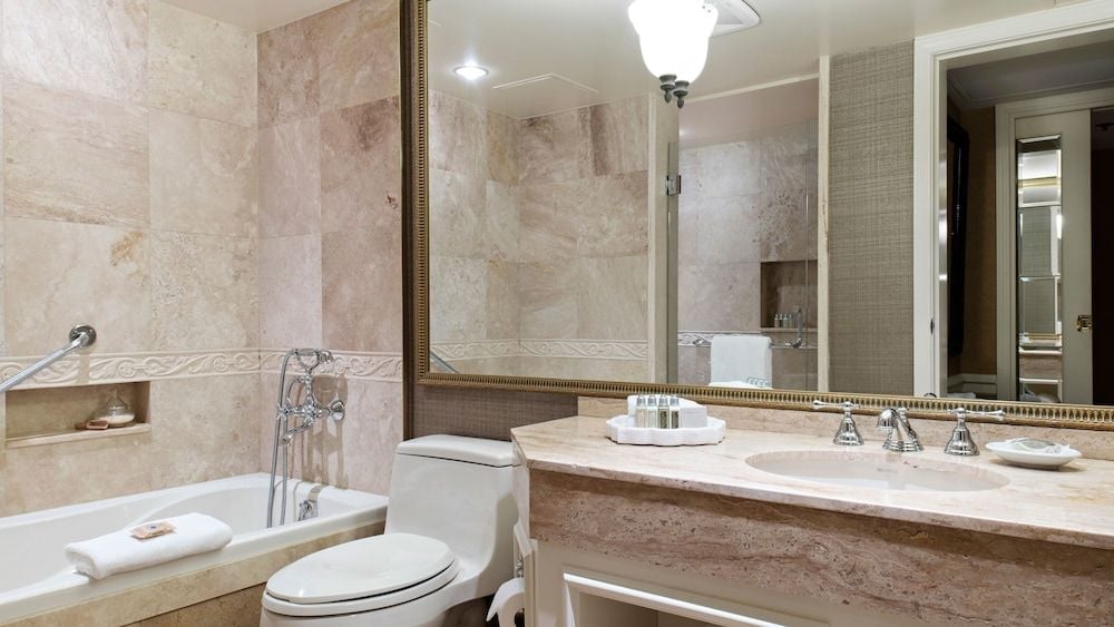 Travertine walls and marble countertops create a spa-like sanctuary, where chrome fixtures gleam against warm stone and fluffy white towels await beside the soaking tub.