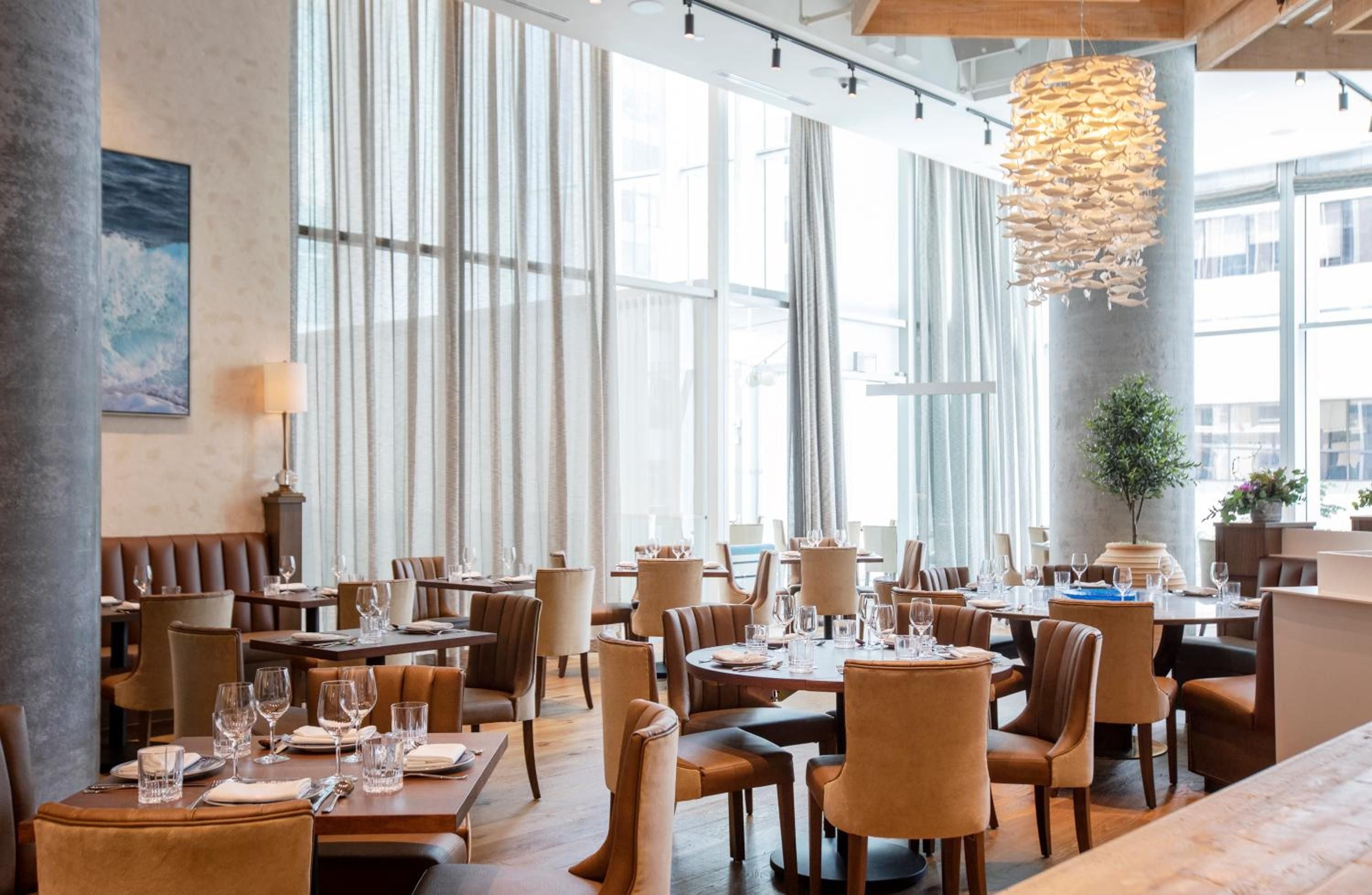 Warm cognac leather chairs and floor-to-ceiling windows create an inviting dining atmosphere, while sculptural pendant lighting adds contemporary flair to this sophisticated restaurant space.