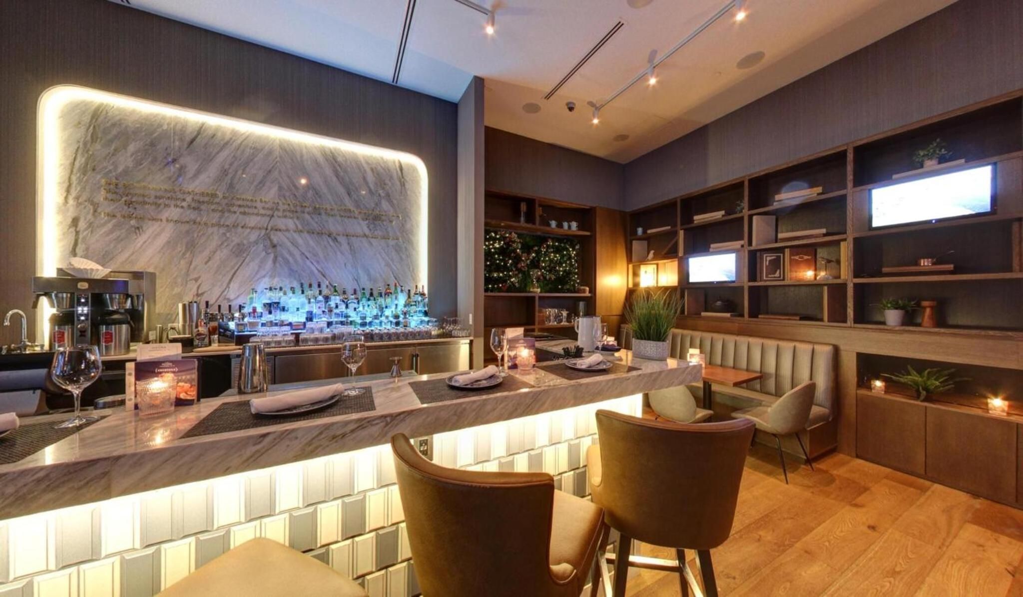 Warm wood tones and illuminated marble create an intimate setting where craft cocktails flow beneath softly lit shelving and contemporary banquette seating.