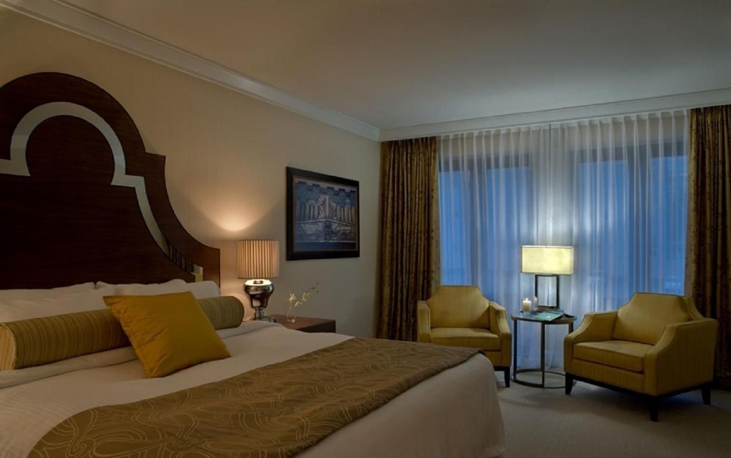 Warm amber lighting bathes the sophisticated guest room, where chocolate leather headboards meet golden upholstery and floor-to-ceiling windows frame the city beyond.
