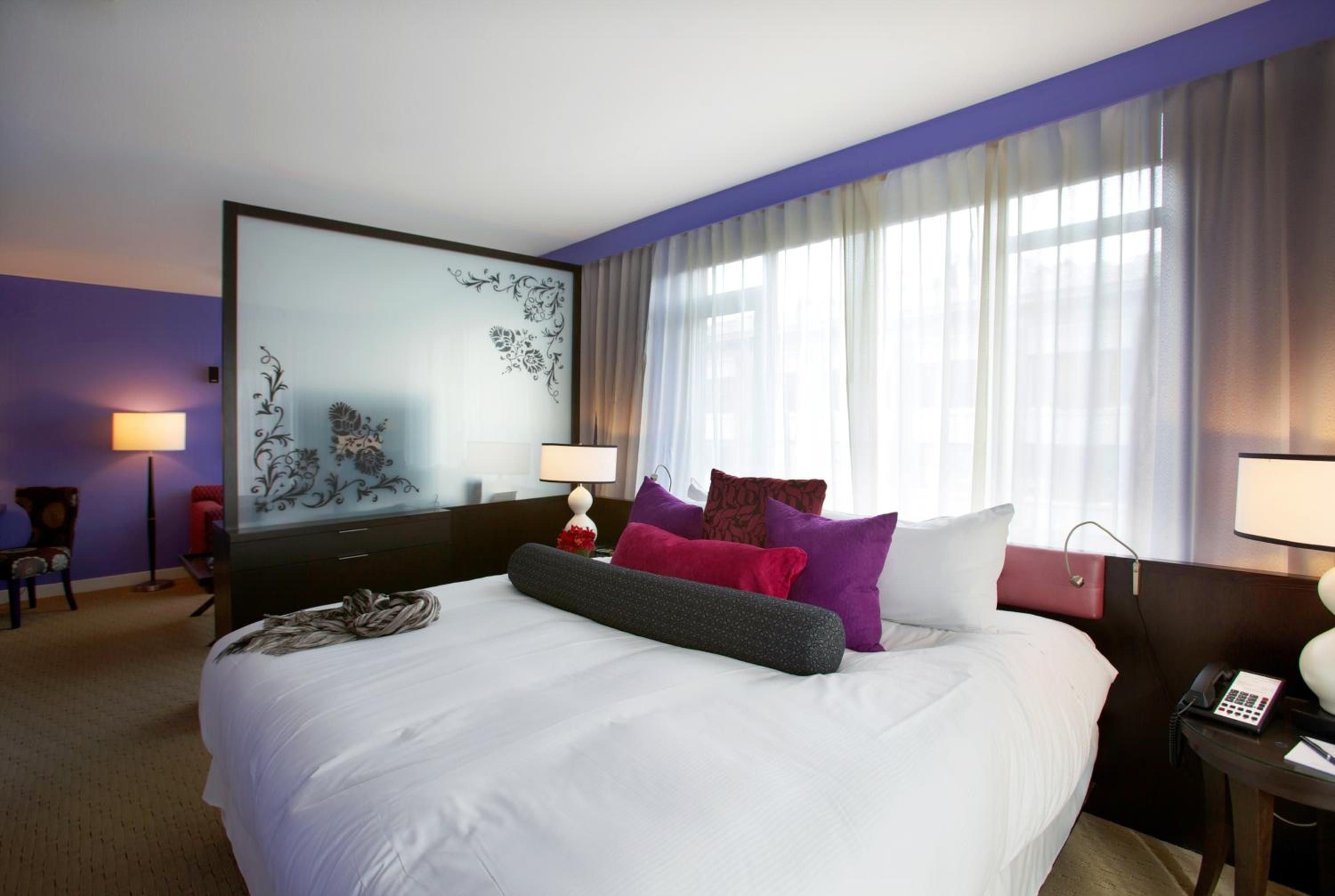 Purple accent walls and jewel-toned pillows create a bold, contemporary sanctuary where natural light filters through flowing curtains onto crisp white linens.