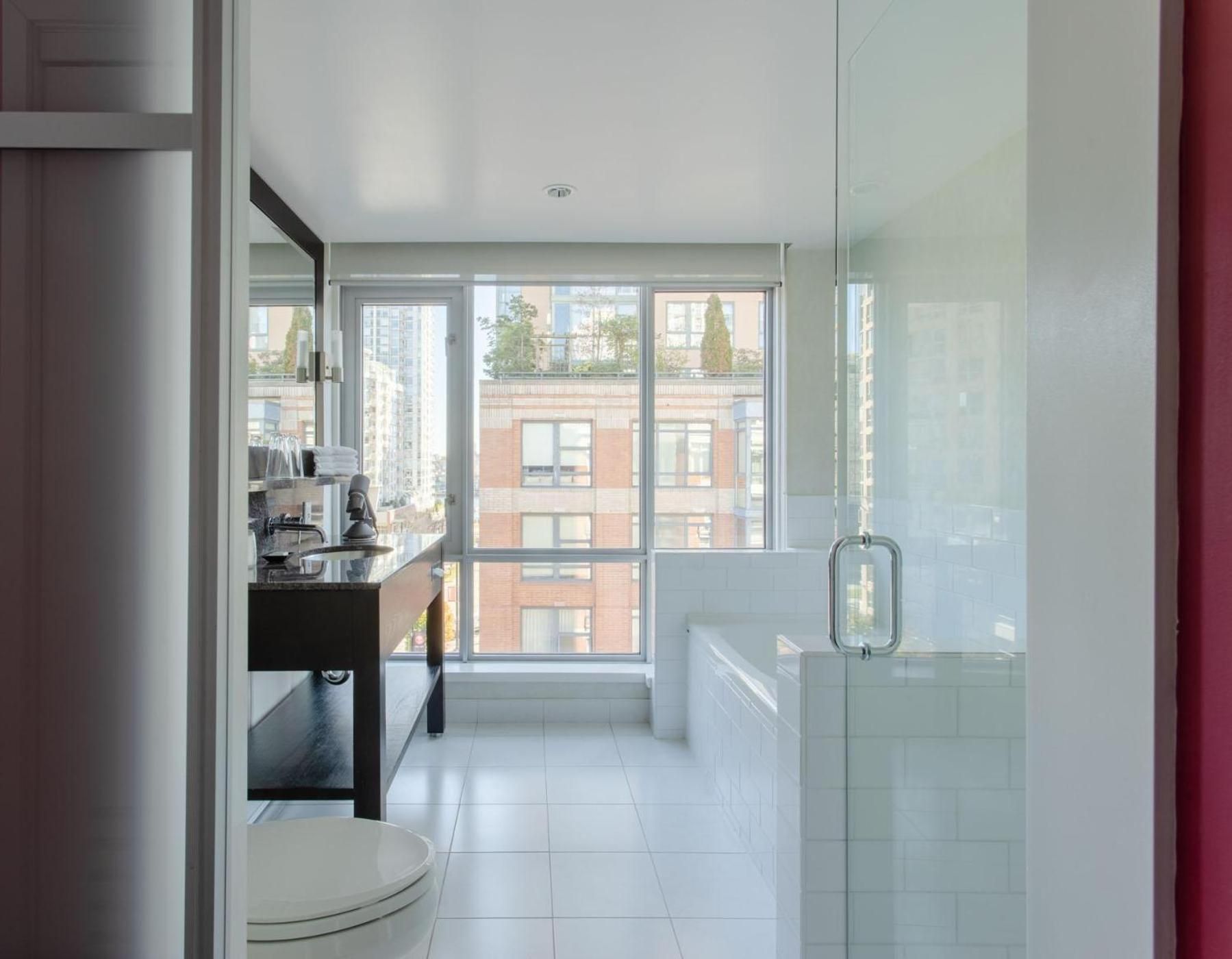 Floor-to-ceiling windows frame red-brick Vancouver rooftops from this sleek bathroom, where marble meets glass in contemporary harmony above pristine white tiles.