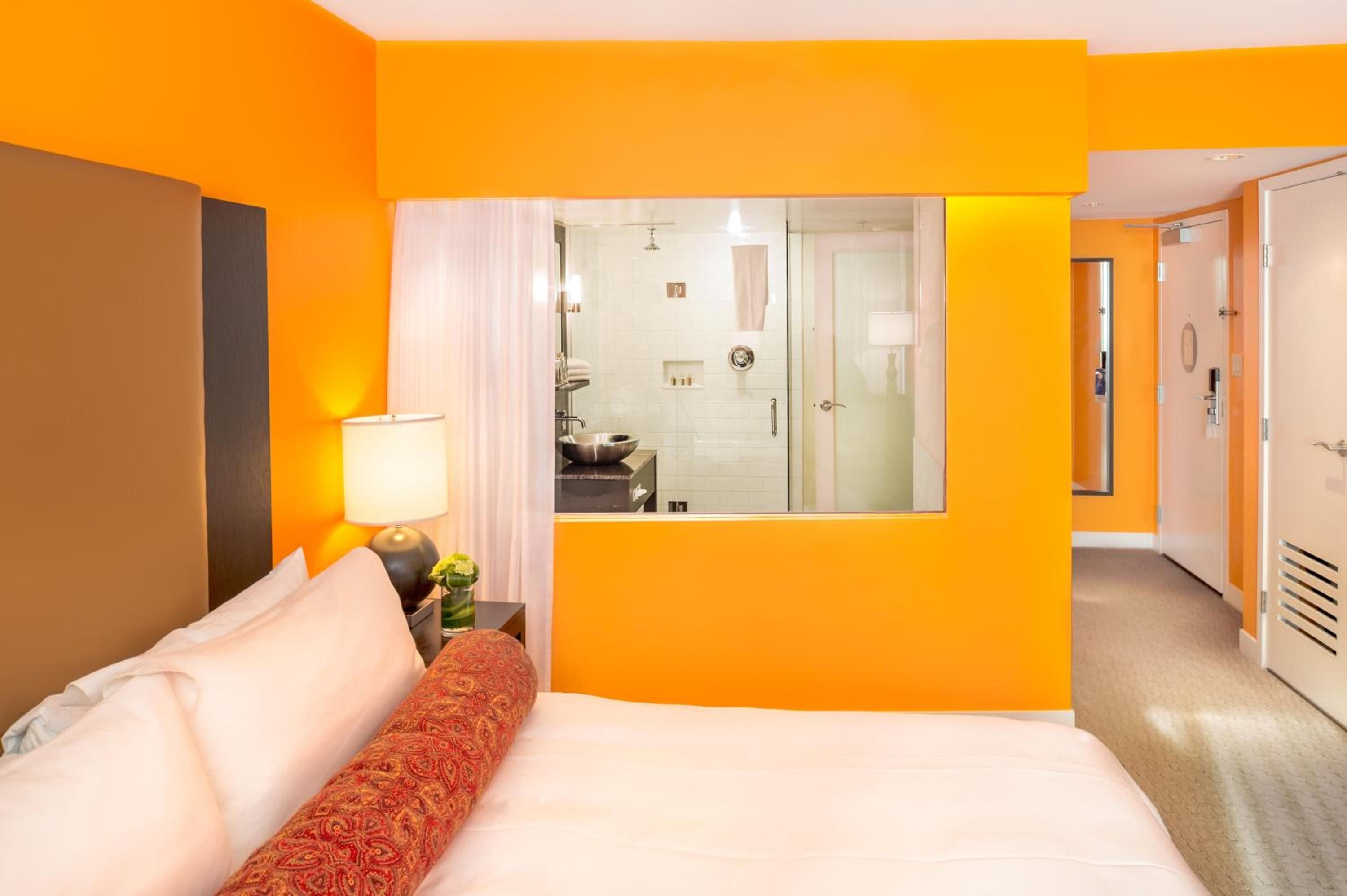 Vibrant orange walls frame an open-concept suite where contemporary sleeping quarters flow seamlessly into a sleek glass-enclosed bathroom with vessel sink and rainfall shower.