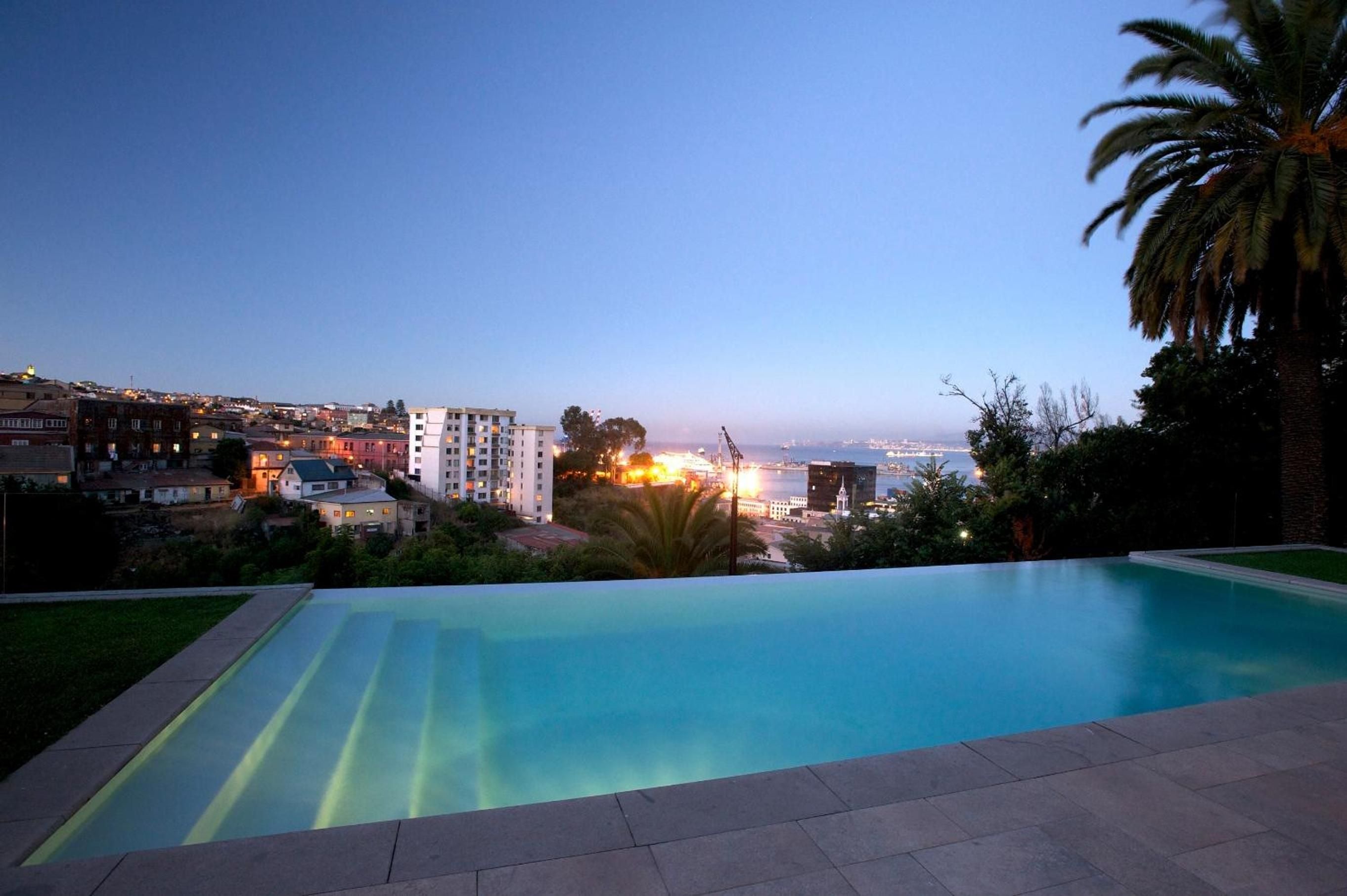 Infinity pool waters glow turquoise against Valparaíso's twinkling hillside lights as twilight paints the Pacific horizon in gentle pastels.