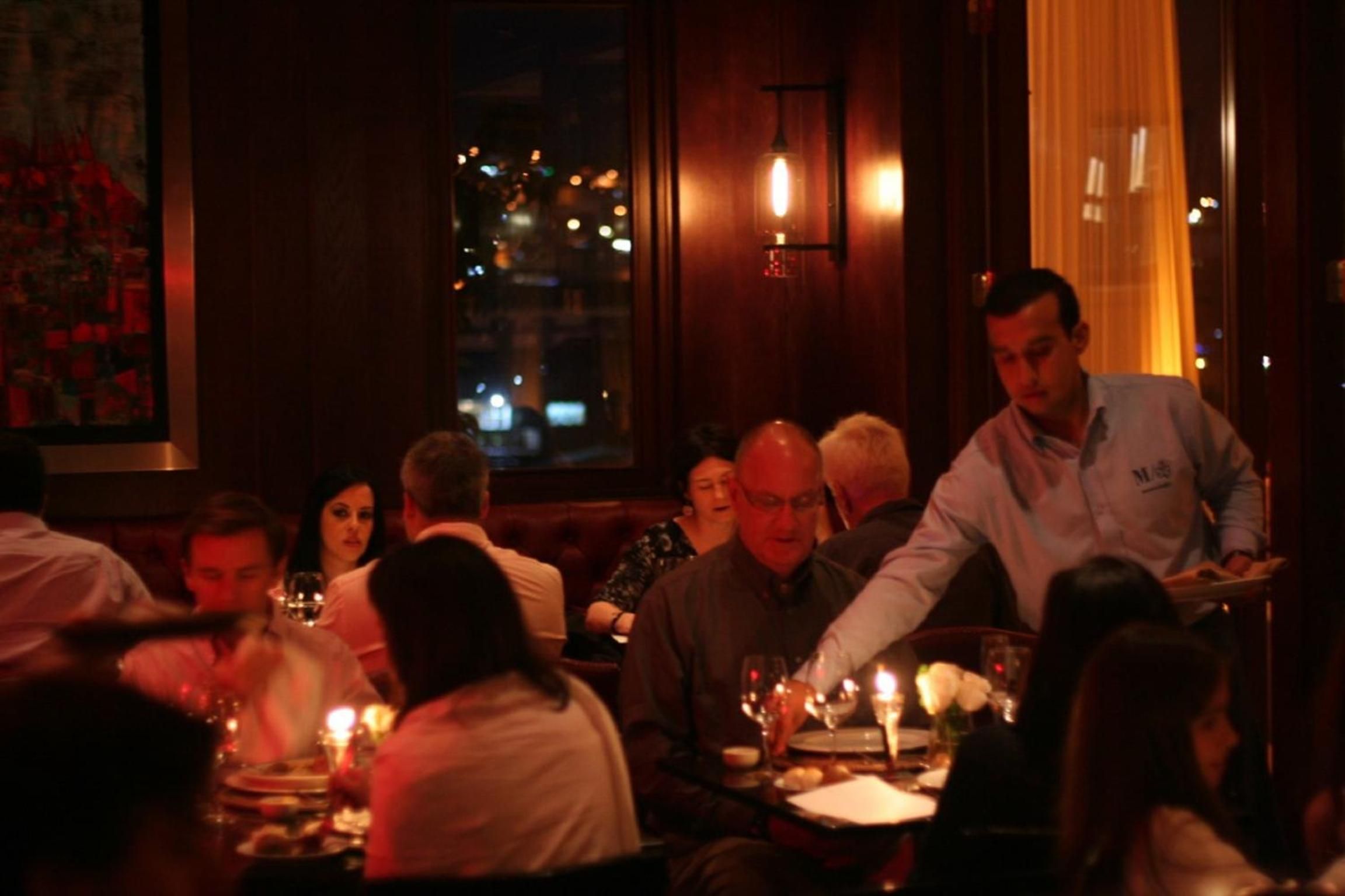 Candlelit tables cast amber glows across intimate conversations while attentive servers glide between guests in this warmly lit dining sanctuary overlooking the harbor lights.