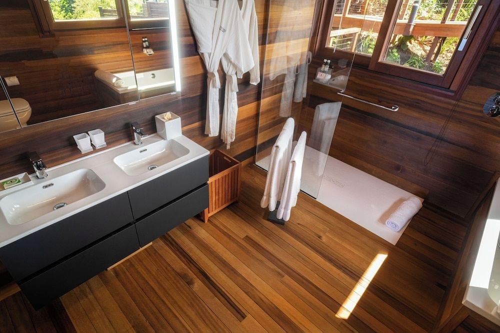 Warm teak envelops this spa-like sanctuary where floating vanities and floor-to-ceiling glass dissolve boundaries between bathing ritual and tropical forest beyond.