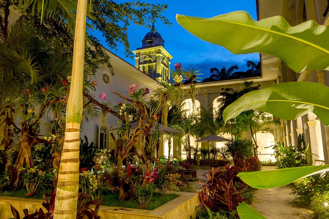 Golden hour illuminates the colonial courtyard where banana palms and tropical blooms frame illuminated archways beneath the historic bell tower's watchful presence.