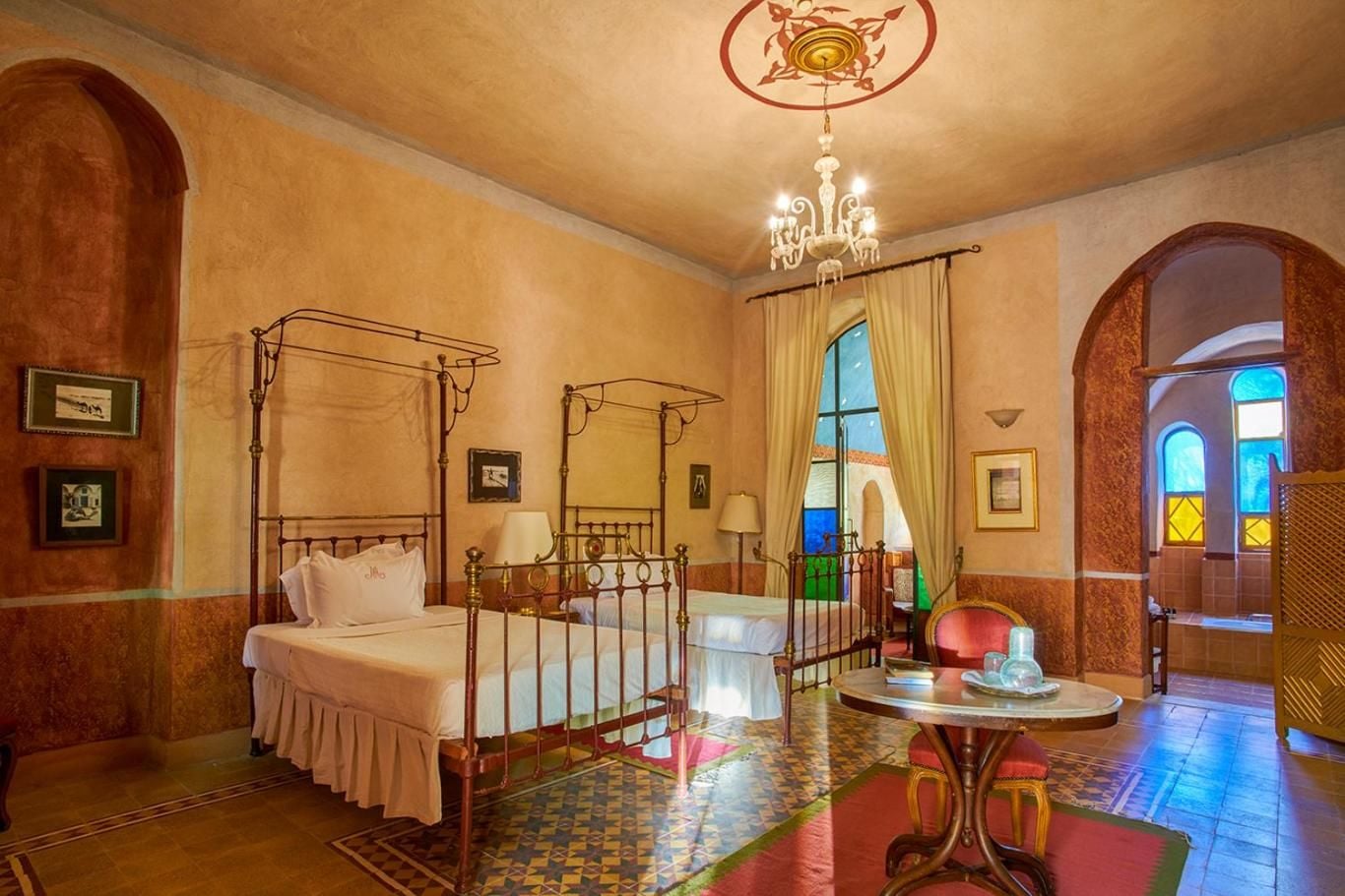 Ornate iron canopy beds and hand-painted ceiling medallions create an intimate Moorish sanctuary, where jewel-toned stained glass bathes terracotta walls in ethereal light.