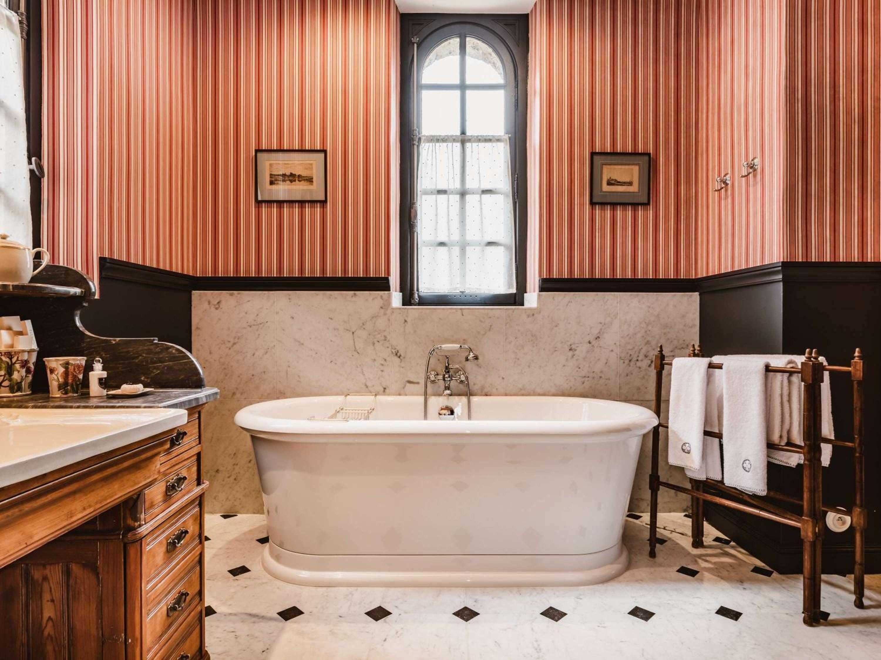 Red-striped wallpaper and marble wainscoting create Victorian elegance in this sumptuous bathroom, where a freestanding tub invites long soaks beneath arched windows.