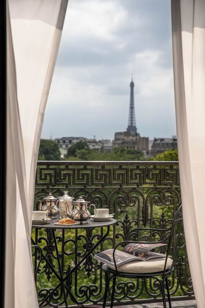 Ornate iron railings frame an enchanting Eiffel Tower vista from this elegant balcony, where silver service and morning pastries await amid billowing curtains.