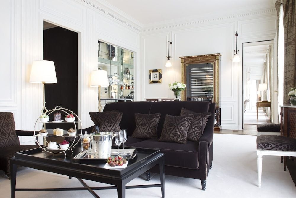 Parisian elegance unfolds in this sophisticated suite where charcoal velvet sofas, gleaming marble surfaces, and crystal champagne service create an intimate sanctuary of refined luxury.