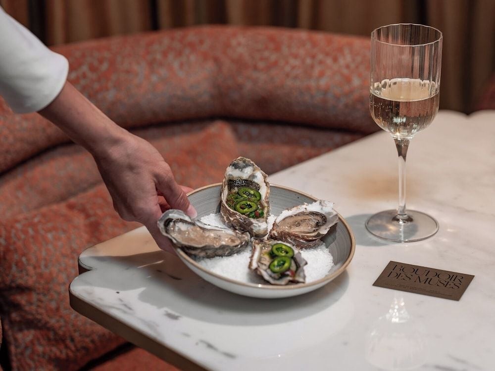 Fresh oysters crowned with jalapeño arrive on crushed ice alongside sparkling wine, creating an intimate culinary moment against coral velvet banquettes.