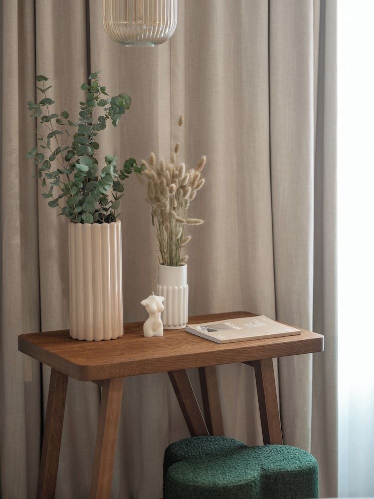 Warm sunlight filters through linen curtains, illuminating a thoughtfully curated vignette where fluted ceramic vessels hold eucalyptus stems above well-worn books and emerald bouclé seating.