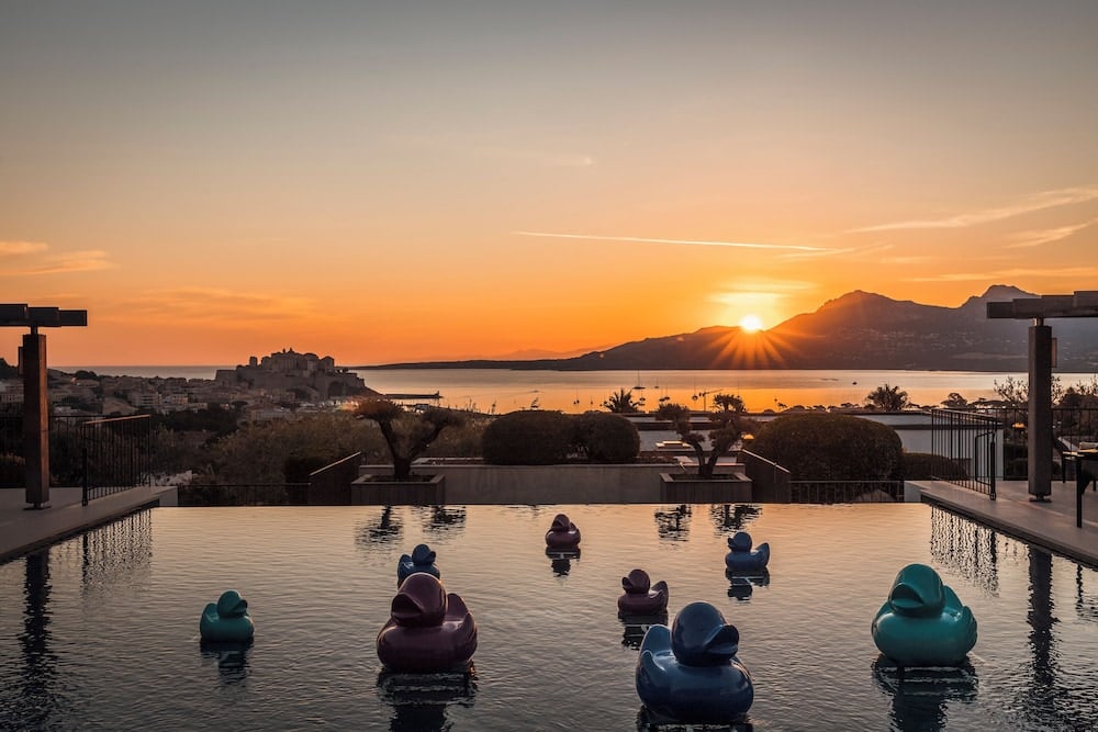 Giant rubber ducks drift across the infinity pool as sunset ignites the Corsican coastline, transforming La Villa Calvi's terrace into a whimsical amphitheater overlooking Calvi's ancient citadel.