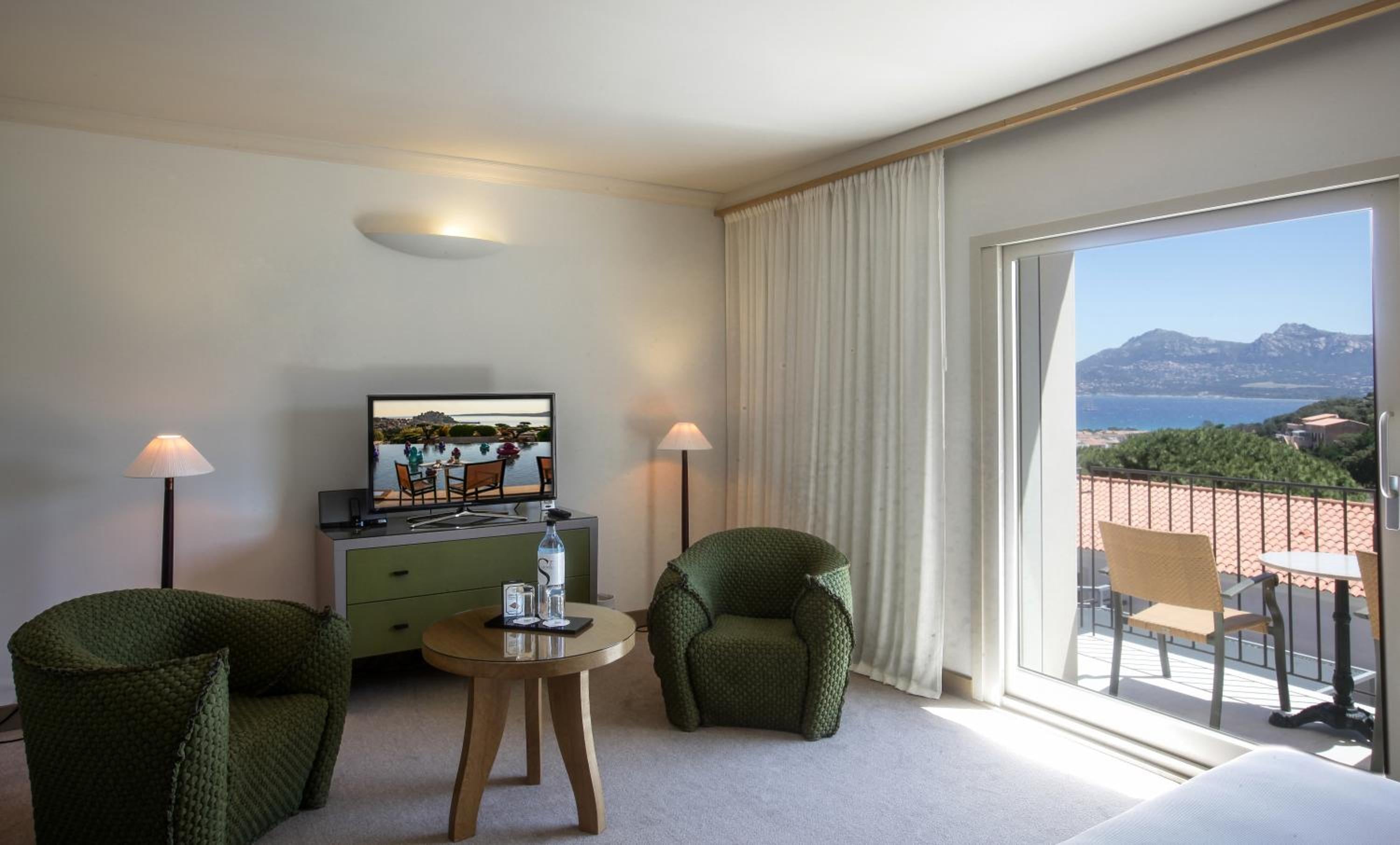 Emerald armchairs frame panoramic vistas of Corsican coastline from this sun-drenched suite, where afternoon light dances across polished surfaces and terra-cotta rooftops.