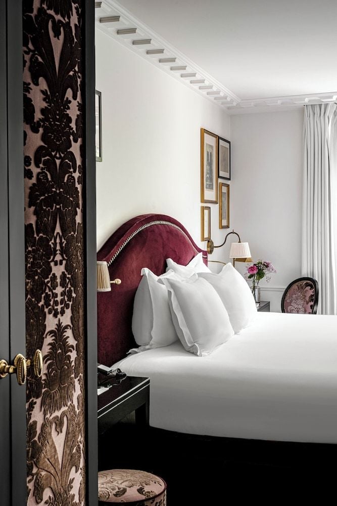 Burgundy velvet headboard and ornate damask wallpaper create intimate French elegance, while crisp white linens and gallery lighting add contemporary refinement to this boutique sanctuary.