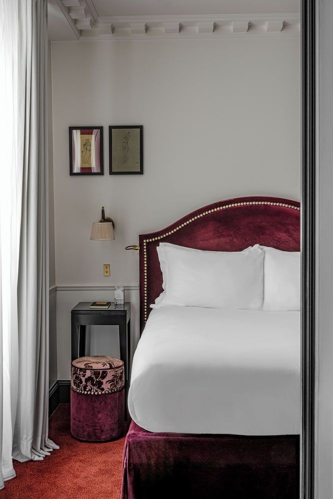 Burgundy velvet headboard with brass studs anchors this intimate guest room, where cream linens meet crimson carpeting beneath coffered ceilings and perfectly curated artwork.