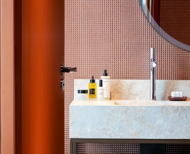 Warm terracotta tones and micro-mosaic tiles create an intimate powder room where luxury skincare products rest on sculptural marble surfaces beneath soft, ambient lighting.