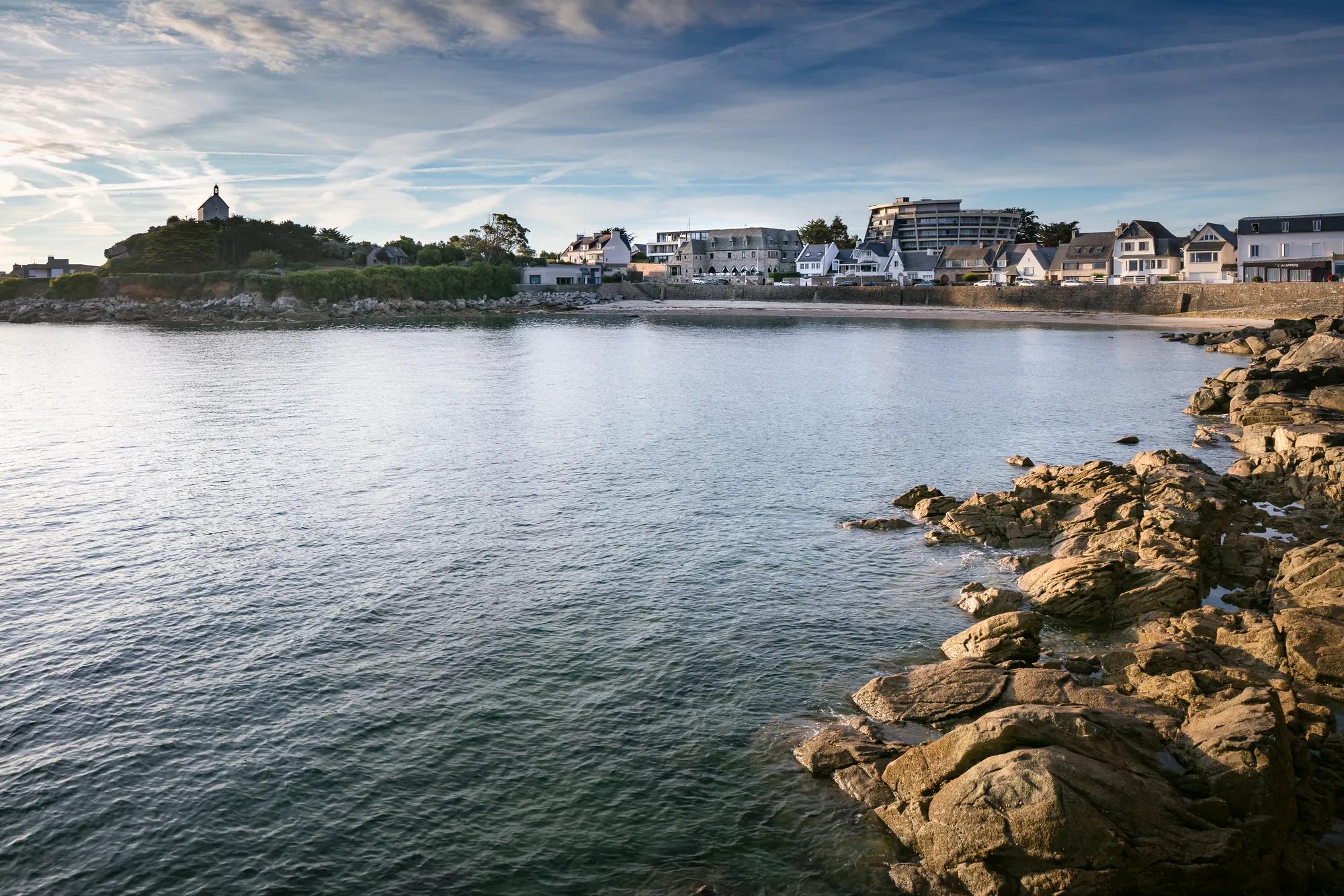 Rocky coastline meets tranquil bay waters where traditional Breton homes and modern seaside architecture create a picturesque maritime tableau under shifting Atlantic clouds.