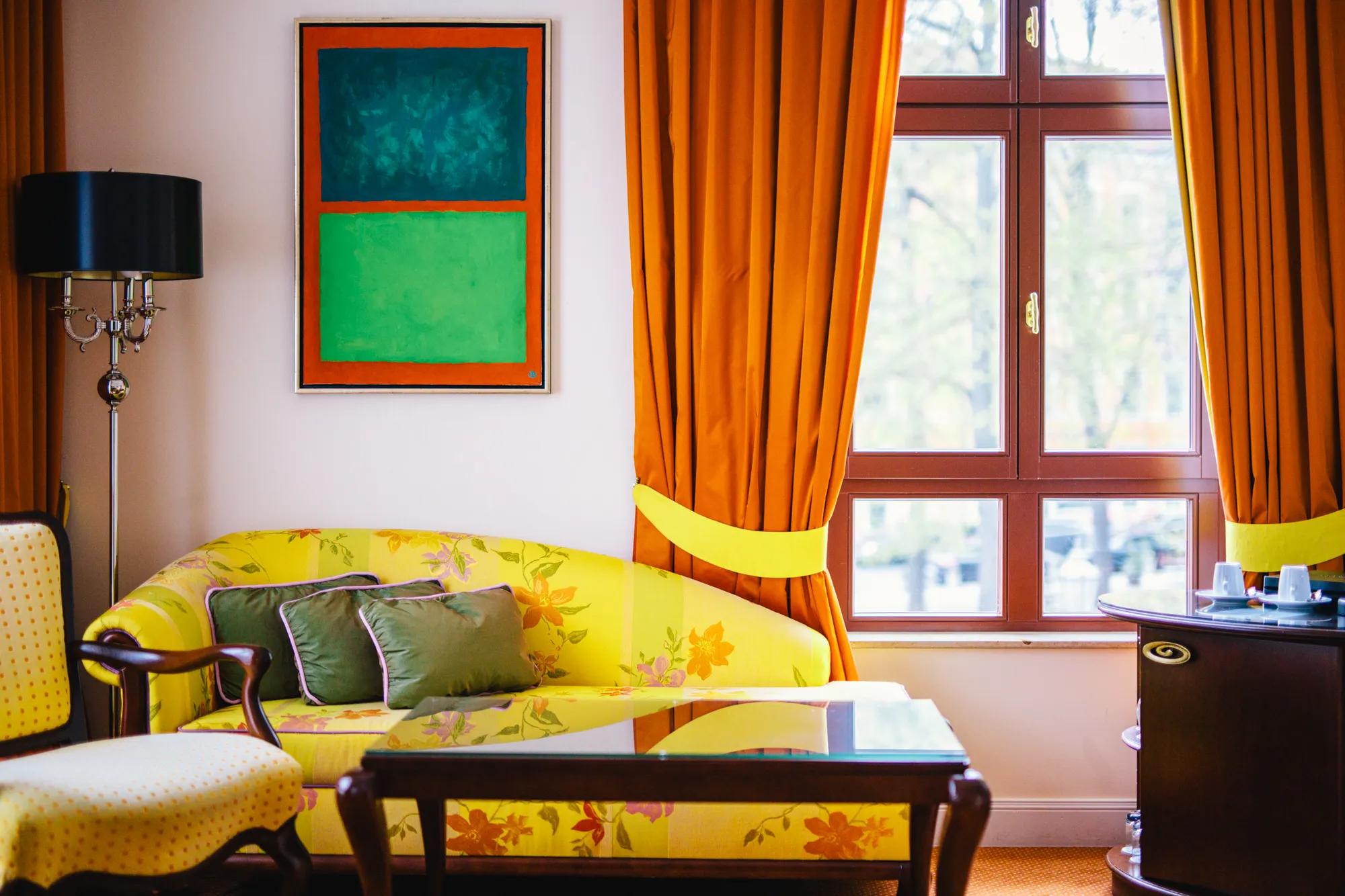 Bold orange curtains frame sunlit windows while a cheerful yellow floral sofa anchors this vibrant sitting area, where contemporary art meets classic European elegance.