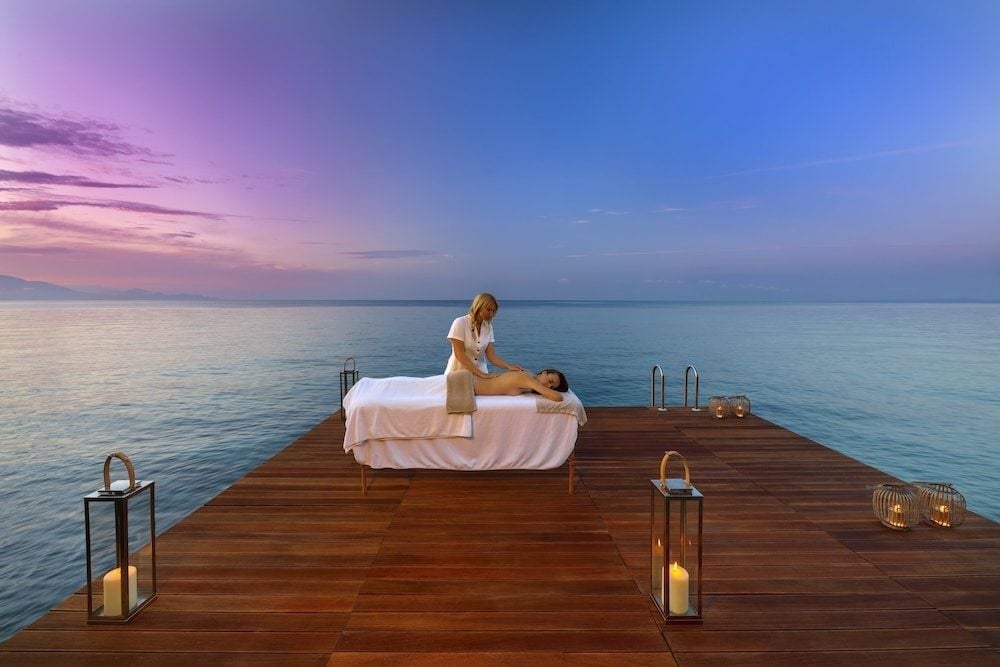 Candlelit massage treatments unfold on a private overwater deck as twilight paints the Ionian Sea in ethereal purples and blues.