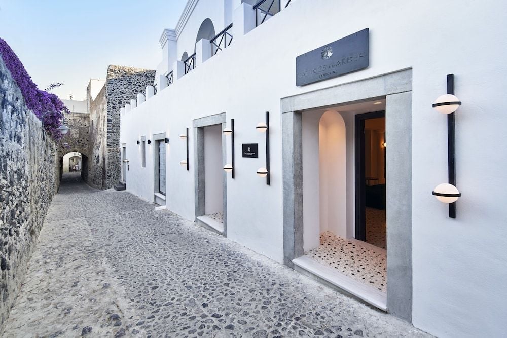 Volcanic stone pathways wind past whitewashed walls and modern lighting fixtures, where traditional Cycladic architecture meets contemporary luxury in this intimate hotel entrance.