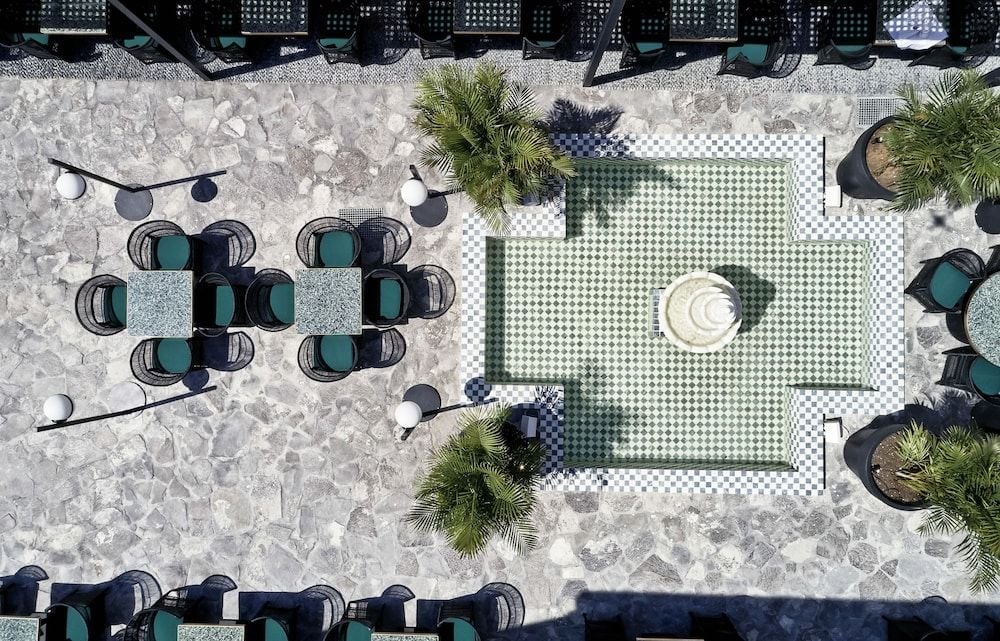 Geometric tiles in soft green create a serene focal point, while palm fronds cast delicate shadows across the meticulously arranged seating areas below.