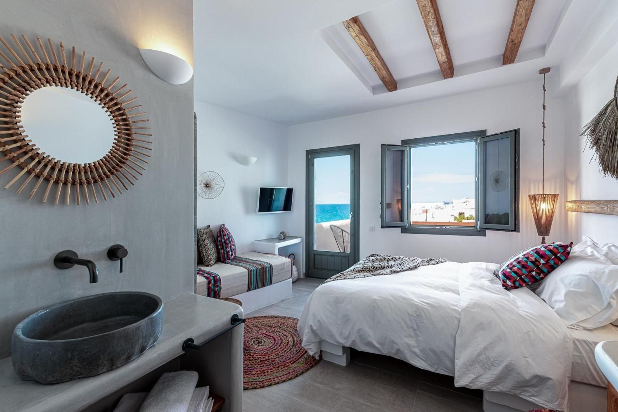 Stone basin and woven textures blend seamlessly in this whitewashed suite, where exposed beams frame azure sea views through traditional Greek shutters.