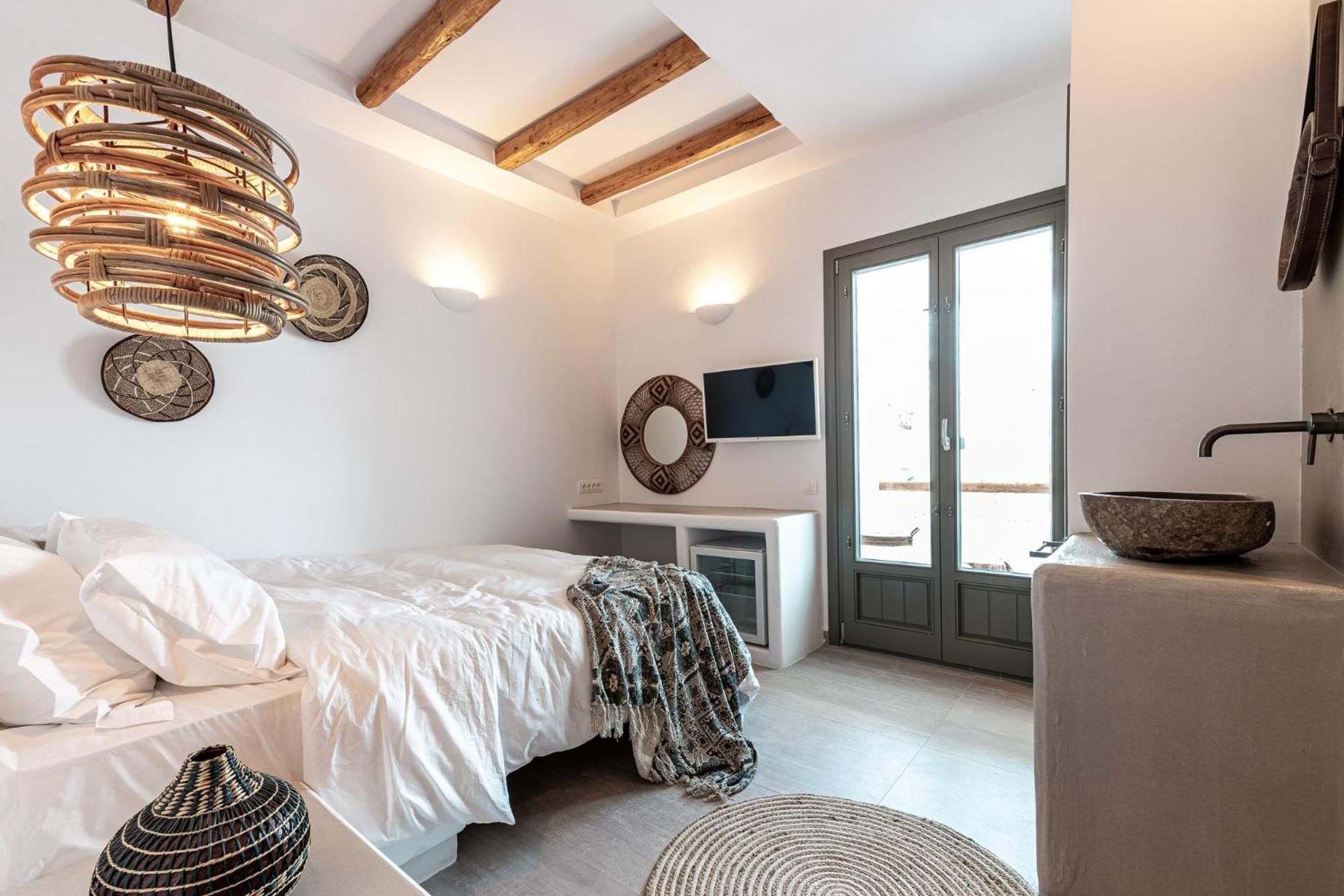 Exposed wooden beams frame crisp white linens while woven pendant lights cast intimate shadows across stone vessels and handcrafted textiles in this serene sanctuary.