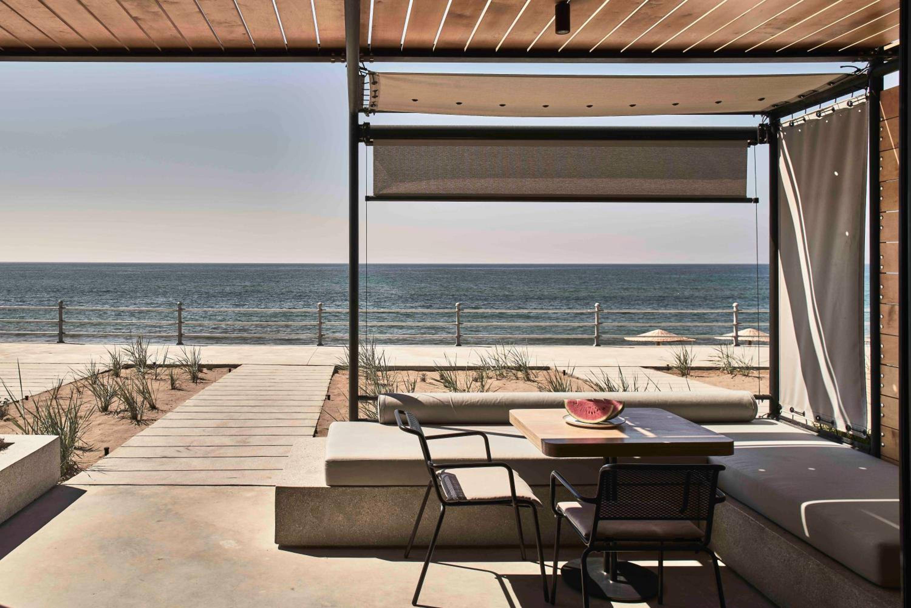 Weathered wood pergolas frame endless Aegean horizons from this beachfront terrace, where industrial-chic furnishings and fresh watermelon create unhurried seaside moments.