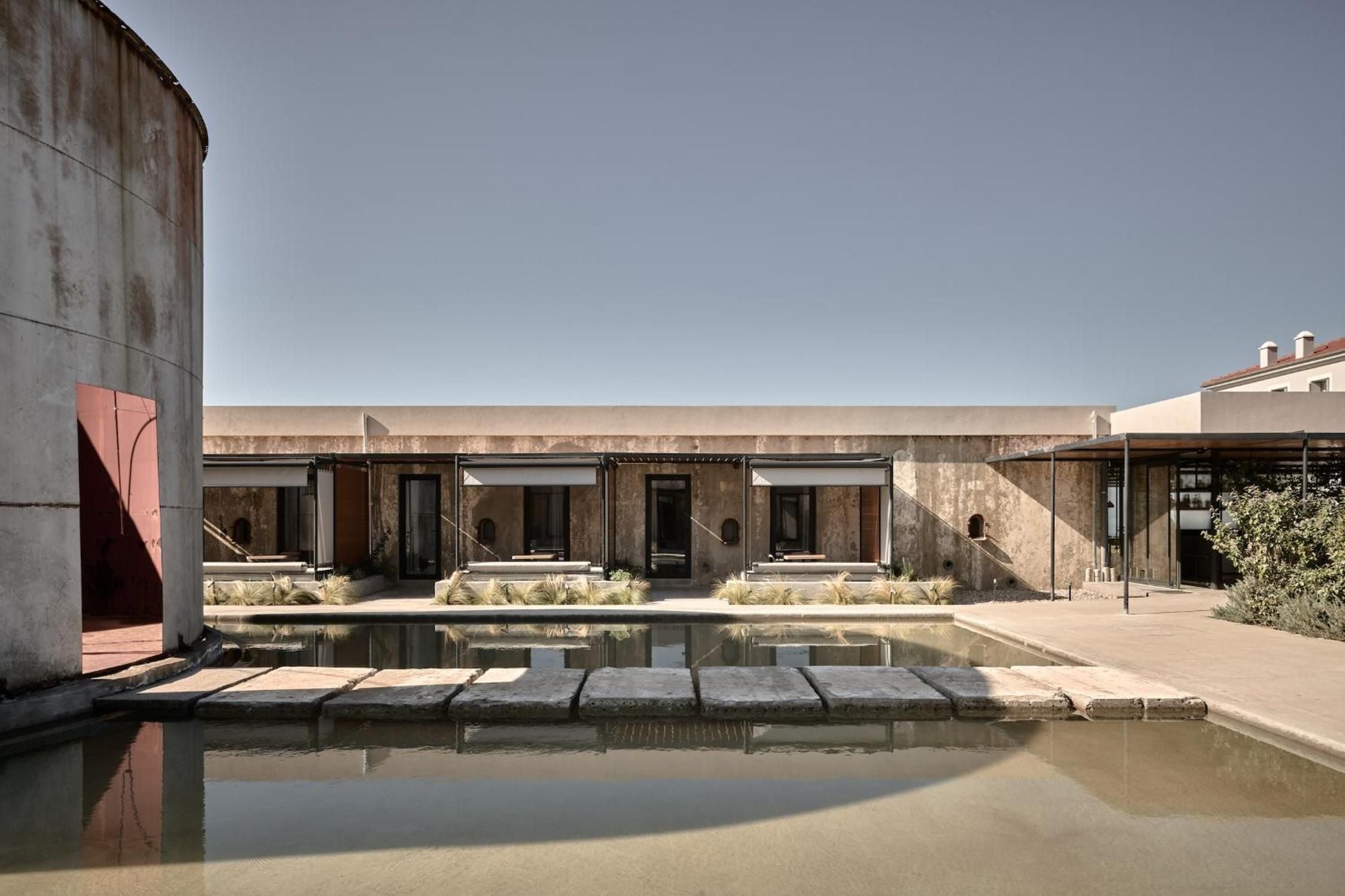 Concrete wine tanks transformed into guest quarters frame a serene reflecting pool, where weathered industrial architecture meets minimalist luxury under Greece's endless sky.