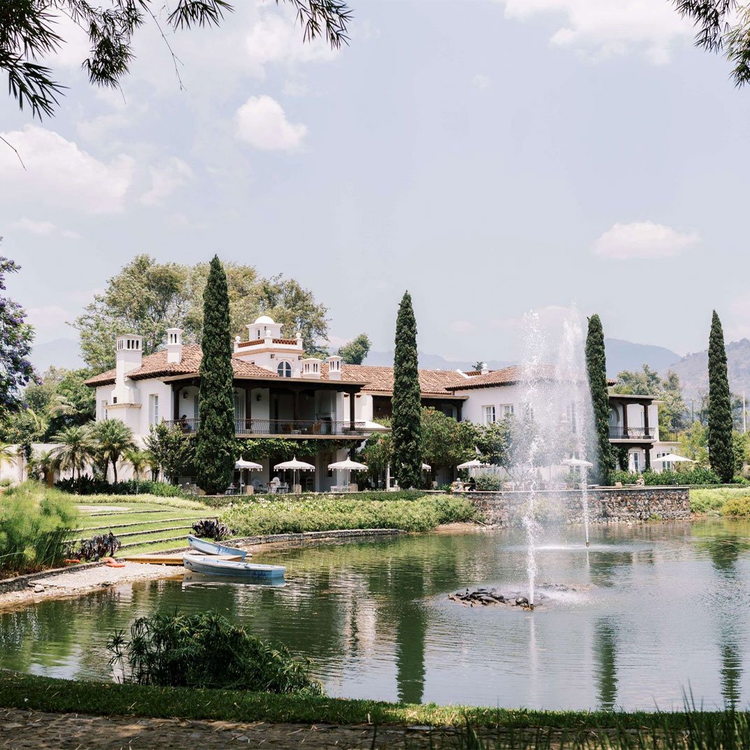 Mediterranean elegance meets tranquil waterside luxury, where cypress trees frame terracotta rooftops and a dancing fountain creates ripples across the serene lagoon.
