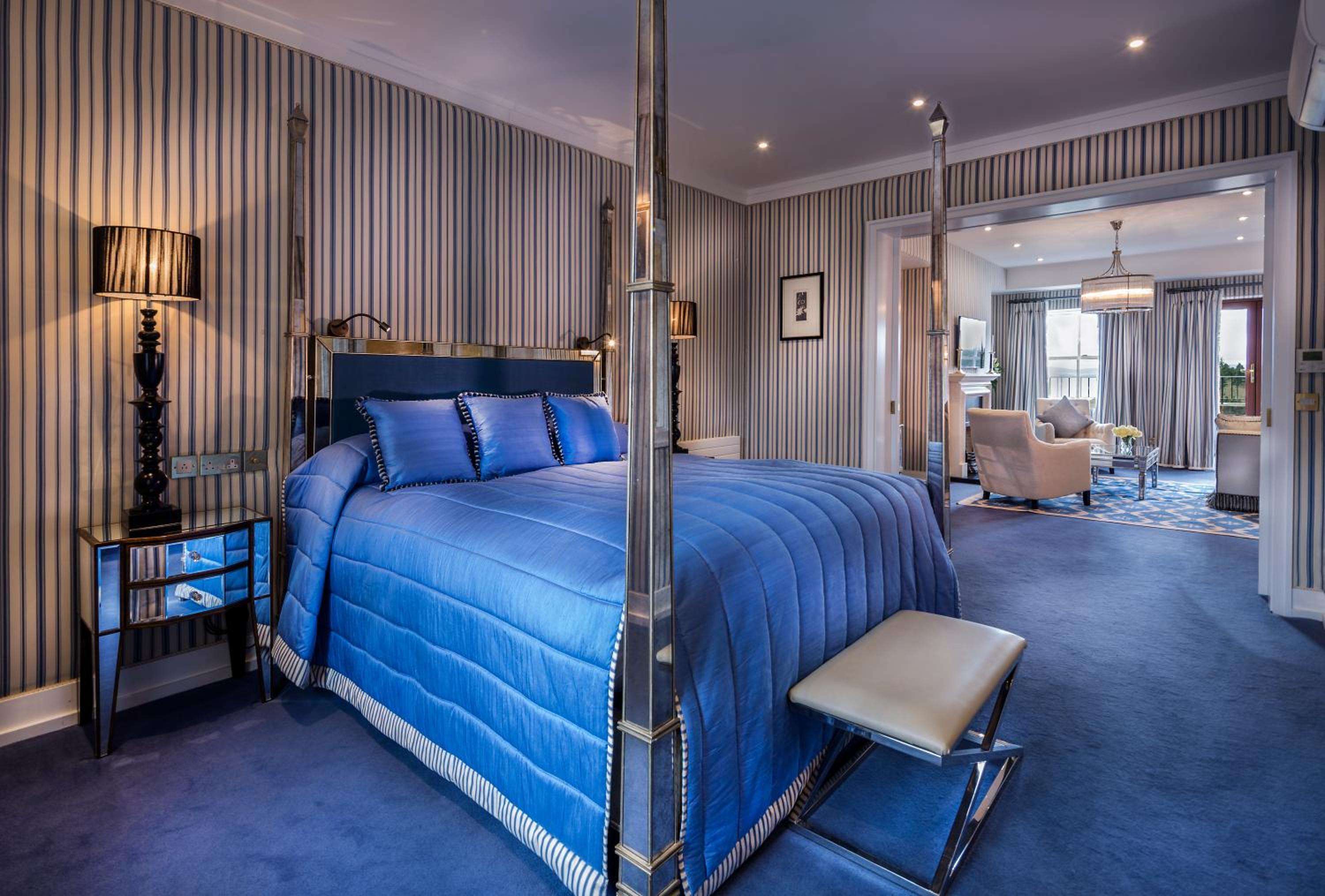 Sapphire silk bedding and polished chrome four-poster frames create regal intimacy in this sumptuous suite, where striped wallpaper adds refined English country house elegance.