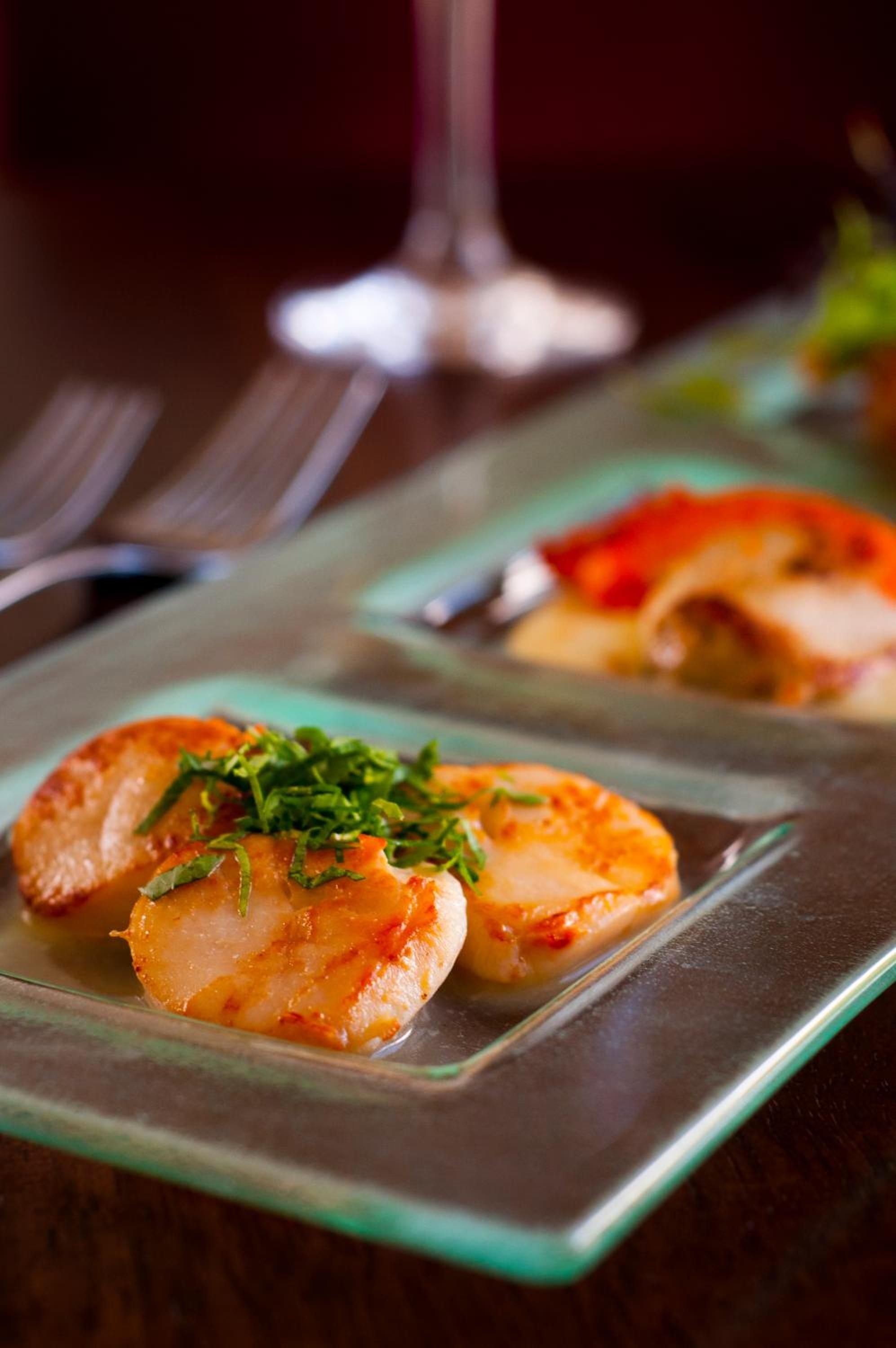 Pan-seared scallops arrive perfectly caramelized on glass plates, their golden surfaces glistening under warm dining room light, finished with bright herbs that promise the chef's meticulous attention.
