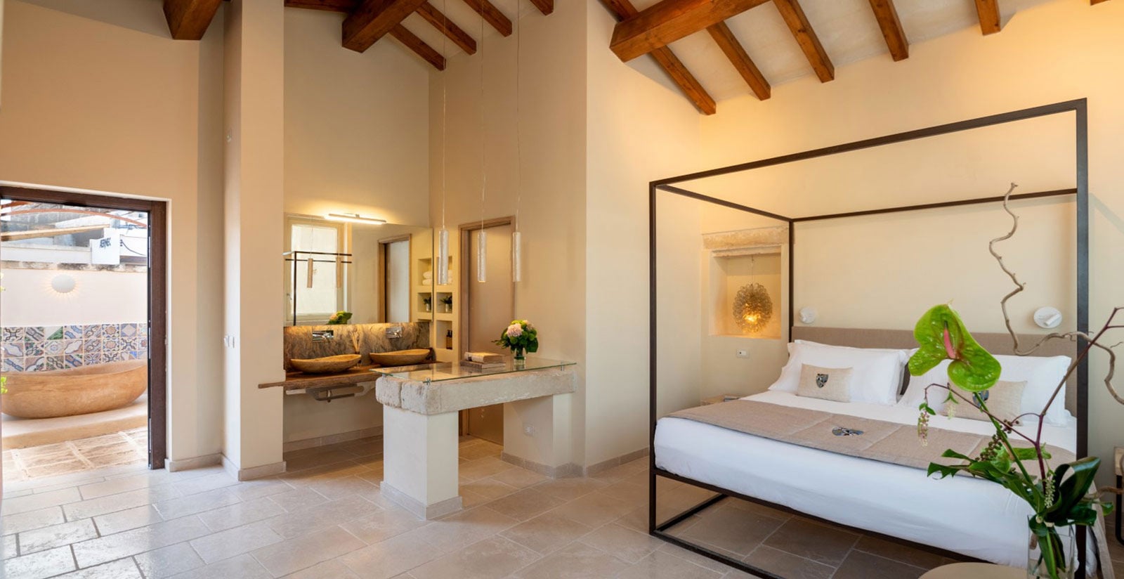 Exposed wooden beams frame this serene suite where natural stone vanities and a sculptural four-poster bed create an intimate sanctuary bathed in golden light.