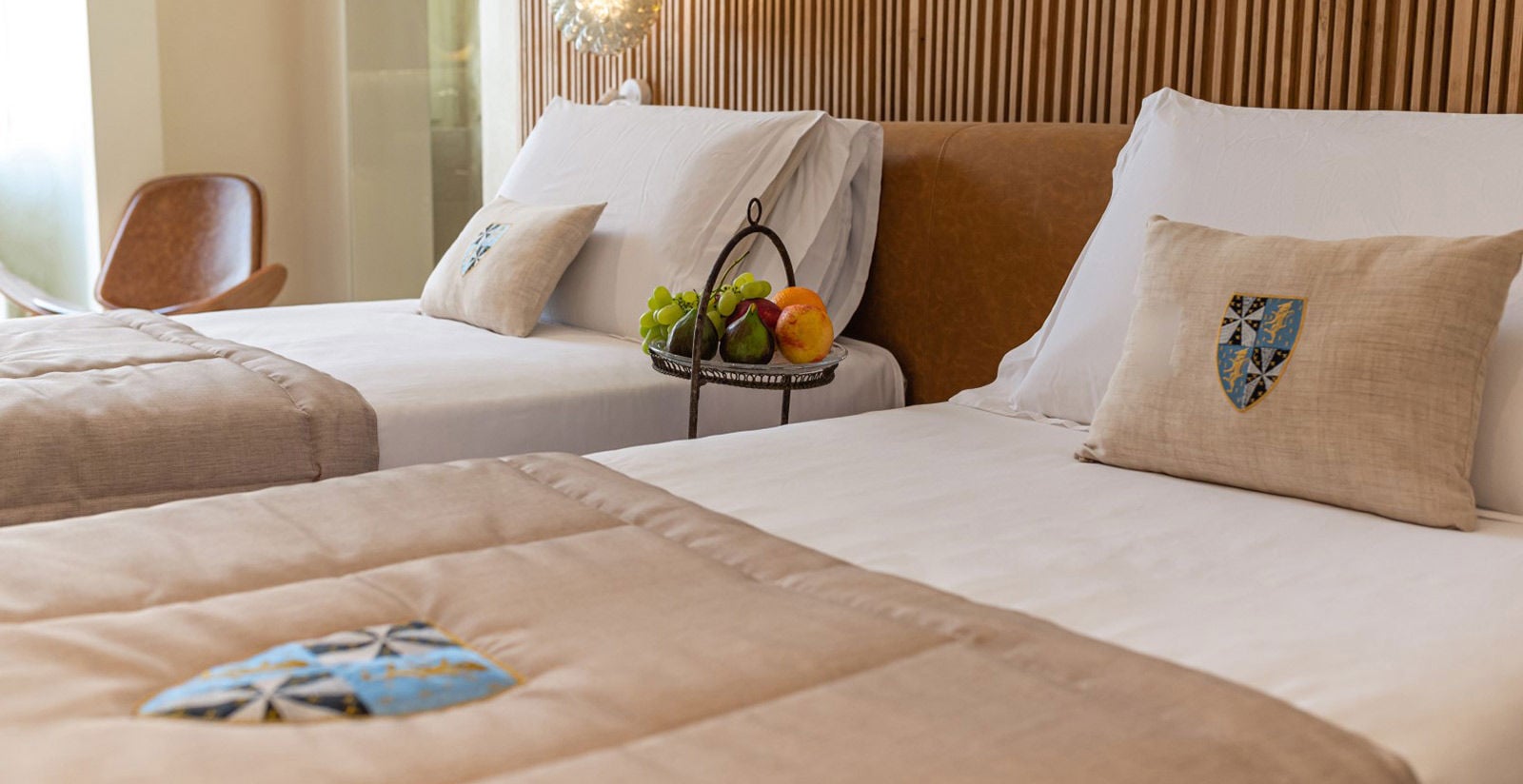 Crisp linens embrace guests in twin beds adorned with heraldic crests, while fresh fruit awaits arrival beneath warm wood paneling and filtered Mediterranean light.
