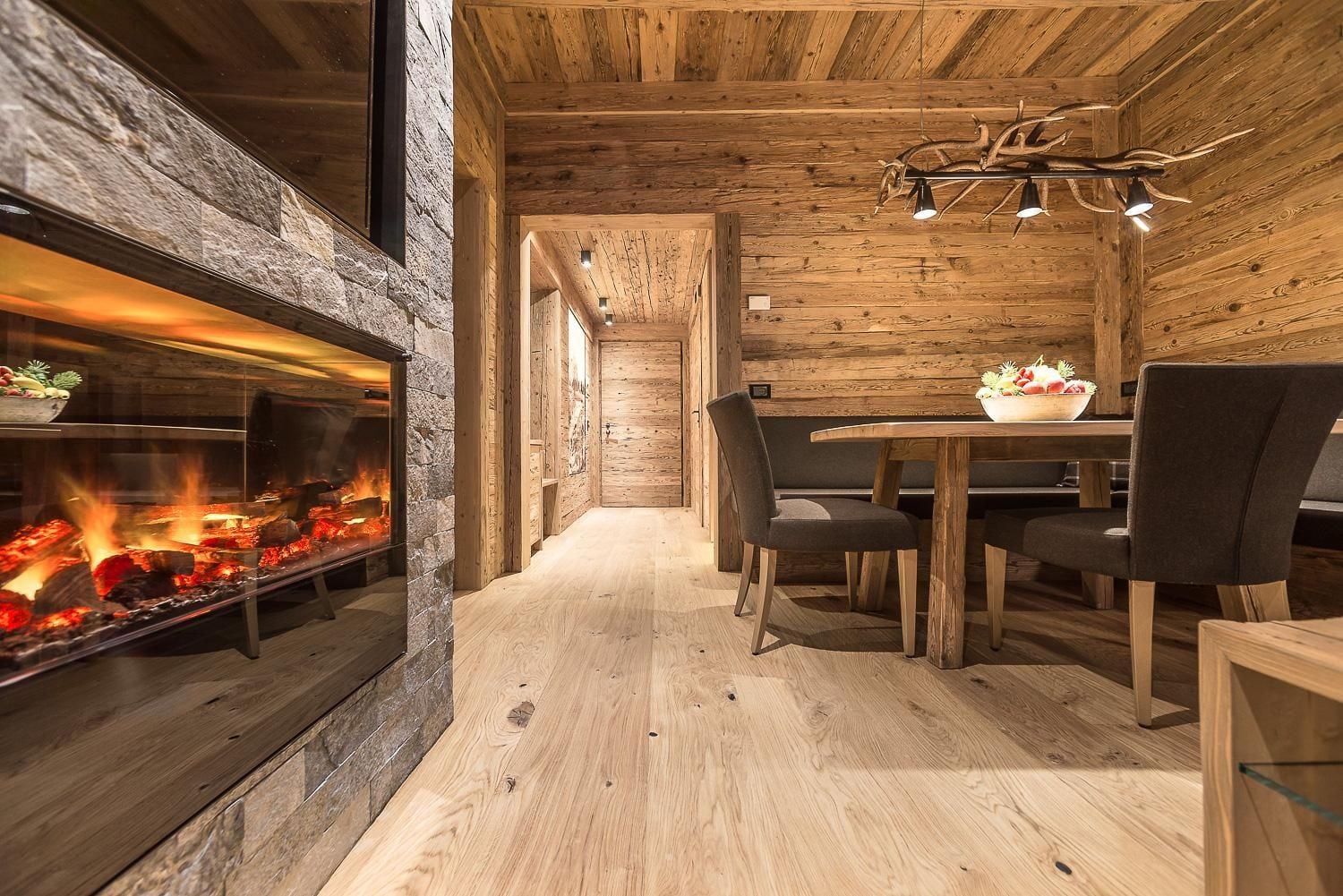 Crackling flames cast amber light across weathered timber walls, where antler chandeliers illuminate intimate dining beneath rustic Alpine beams.