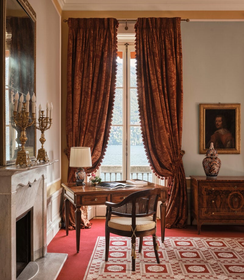 Burnished copper curtains frame lake views in an elegant writing nook, where gilded candelabras and Persian rugs create an atmosphere of old-world refinement.