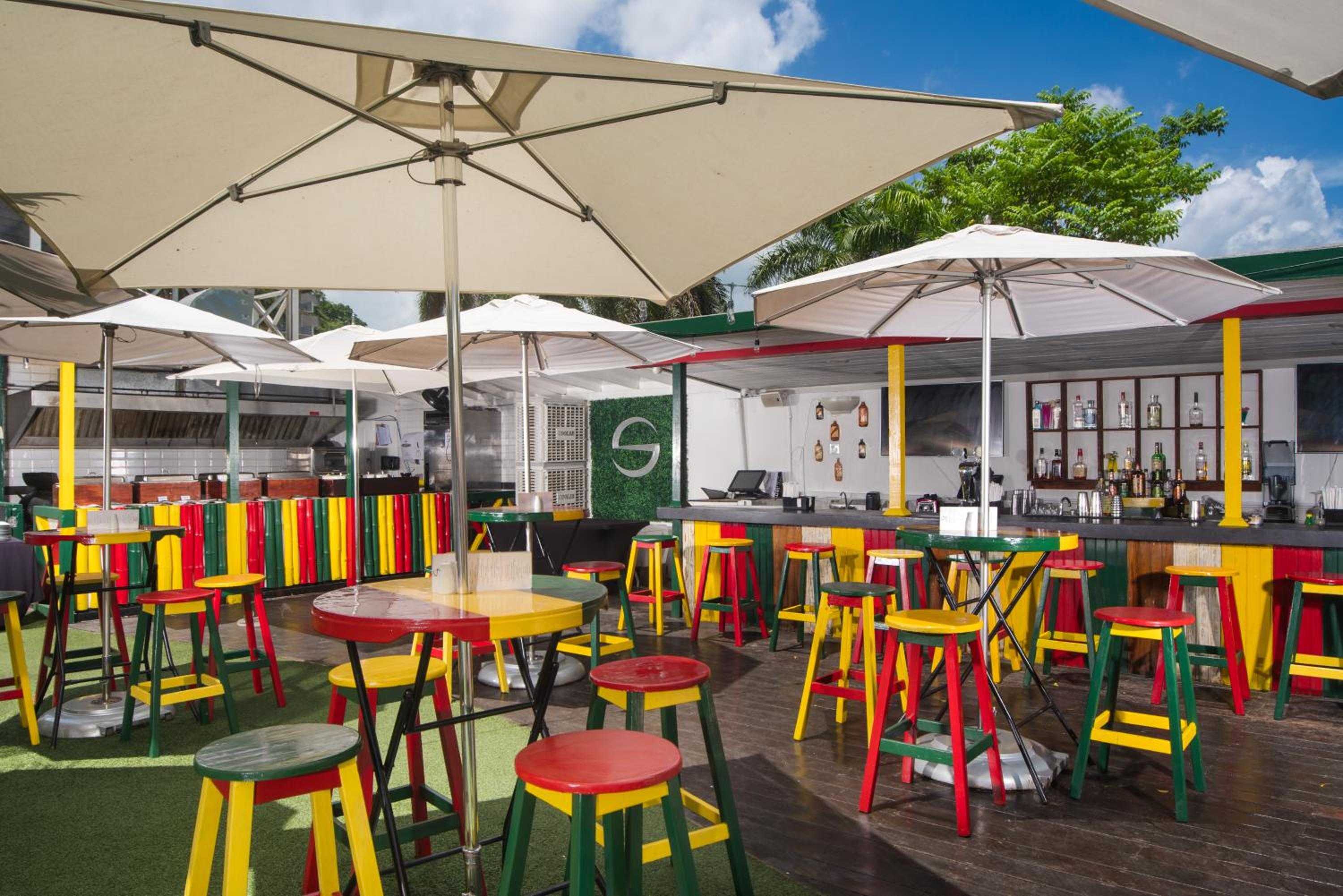 Vibrant Rastafarian-inspired bar stools in red, yellow, and green create a spirited reggae vibe under crisp white umbrellas, where tropical cocktails flow freely.