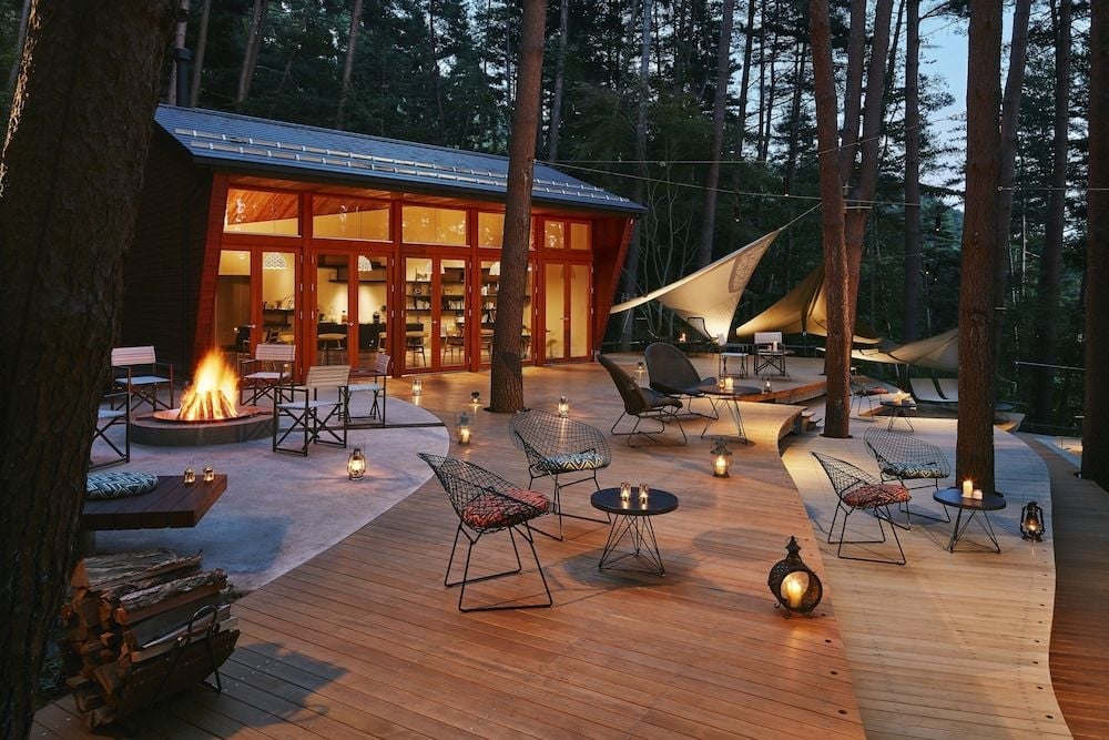 Lantern-lit wooden decks weave between towering pines, where crackling fire pits and modern wire chairs create an enchanting forest retreat at dusk.