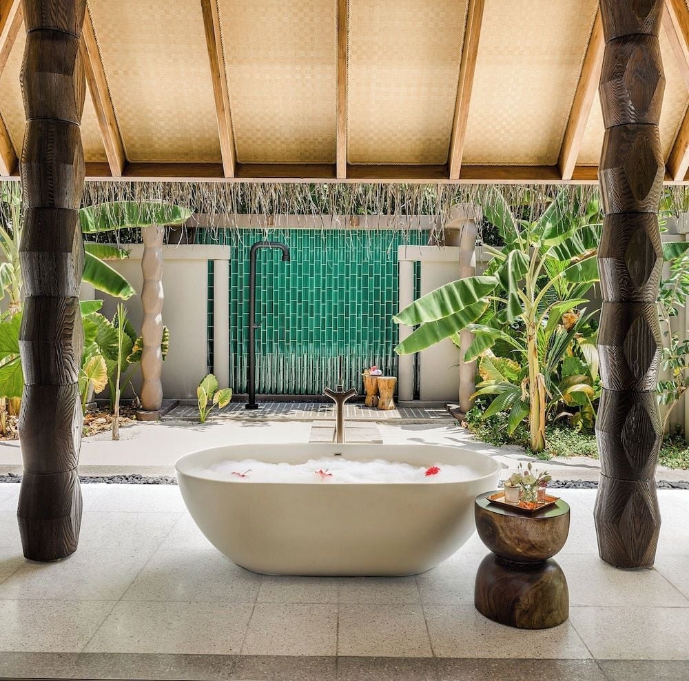 Carved wooden columns frame an outdoor bathing pavilion where emerald tiles shimmer behind a sculptural soaking tub adorned with floating frangipani petals.