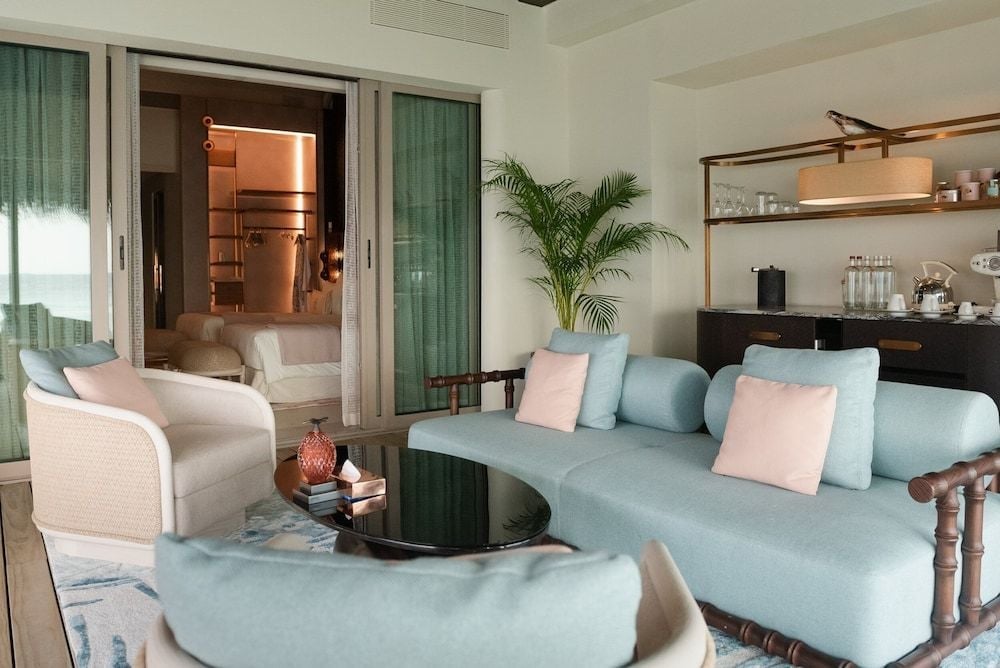 Soft seafoam furnishings and coral-pink accents create a serene sanctuary, where marble surfaces catch tropical light filtering through jade-green curtains into this overwater villa.